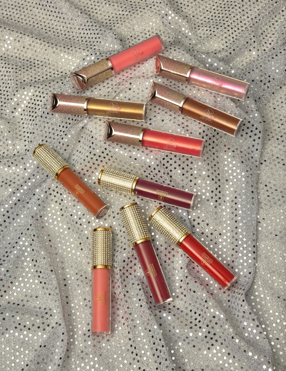 Collection of lip gloss tubes with gold caps, arranged on a sparkly silver fabric background.
