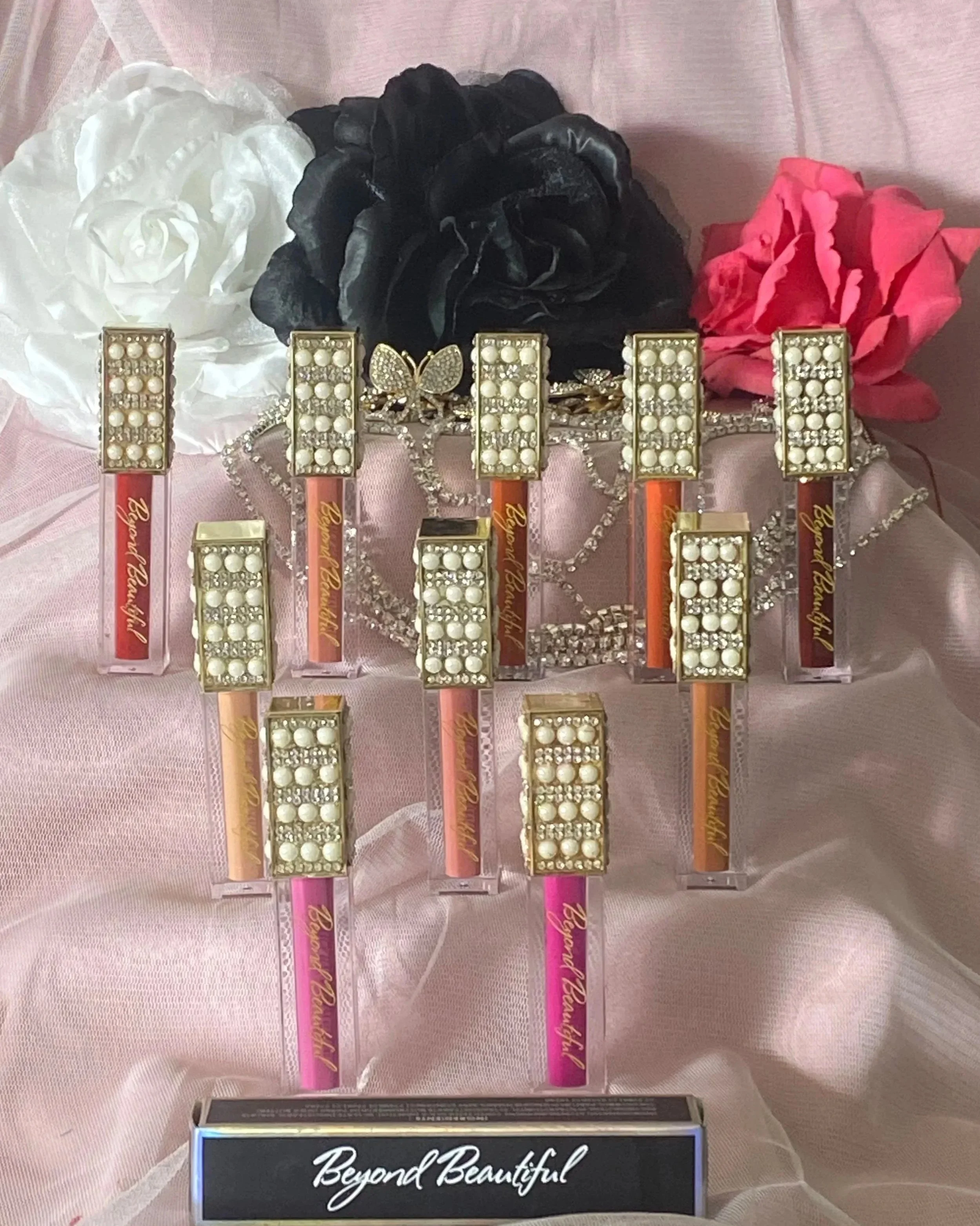 Display of matte lipstick adorned with pearl and rhinestone embellishments, arranged on a pink satin cloth with black, white, and pink artificial flowers in the background, and a sign that reads 'Beyond Beautiful' in front.