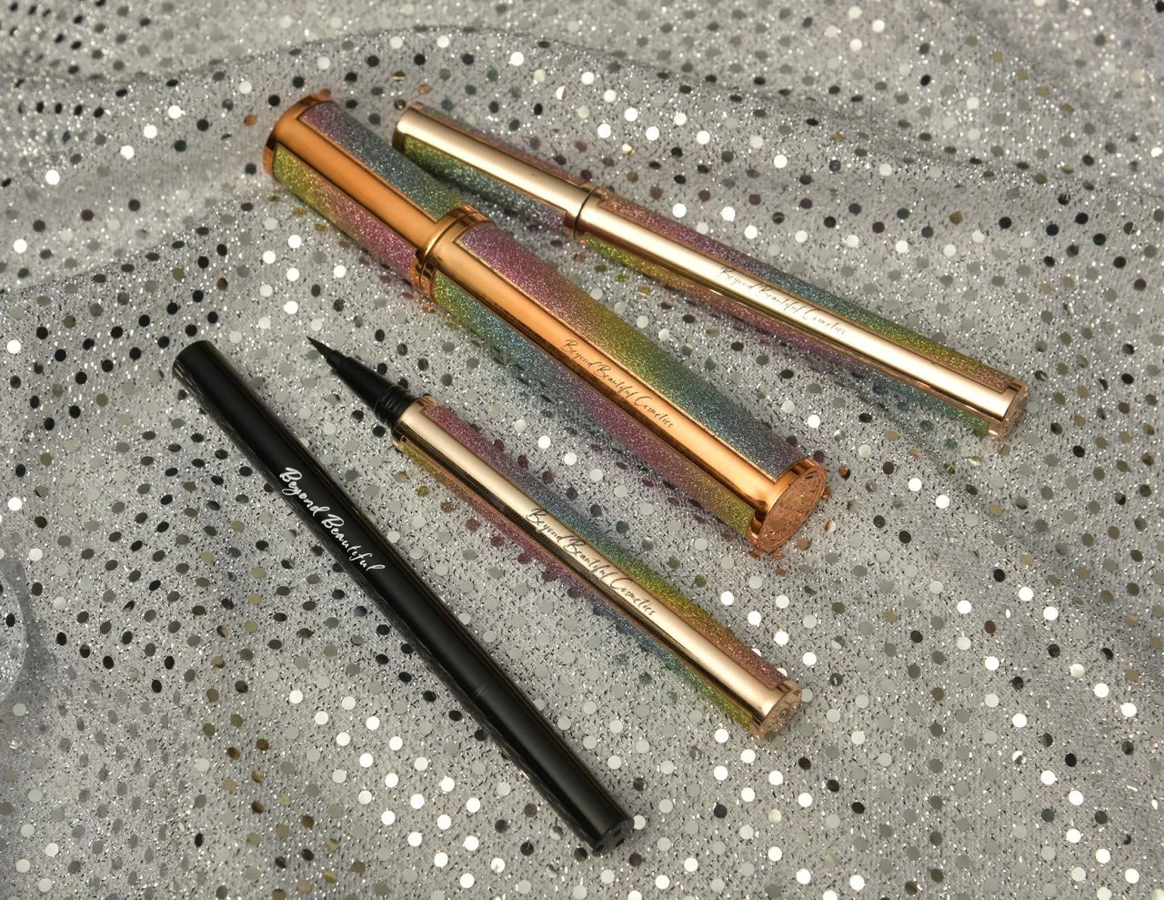 Three glittery, multicolored beauty pens with rose gold accents and one plain black pen, all laid on a sparkly, silver, sequin-covered fabric surface.