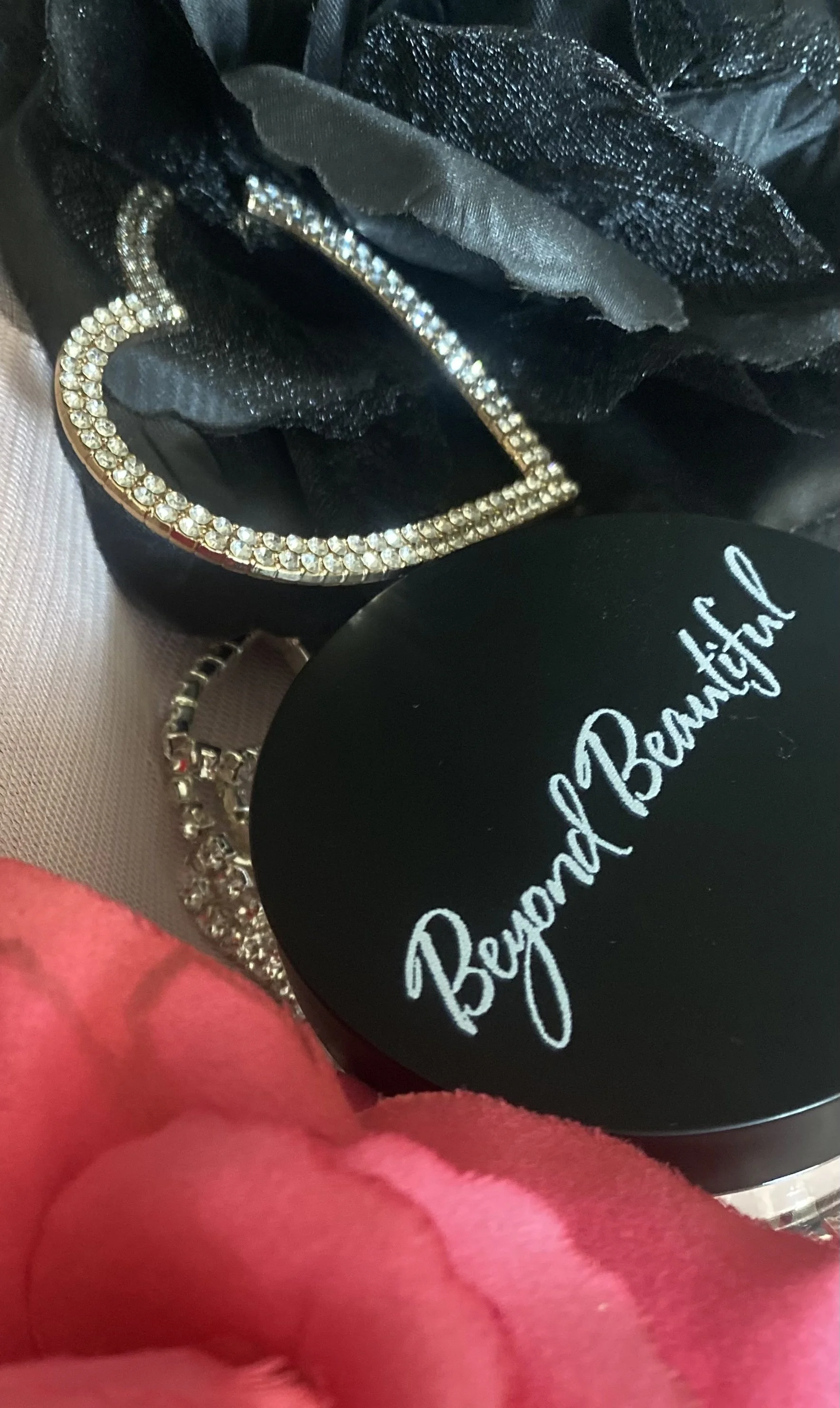 Close-up of jewelry, including a rhinestone-studded heart-shaped bracelet and a diamond bracelet, with black and pink fabric in the background and a round black container labeled 'Beyond Beautiful'. Brand Logo