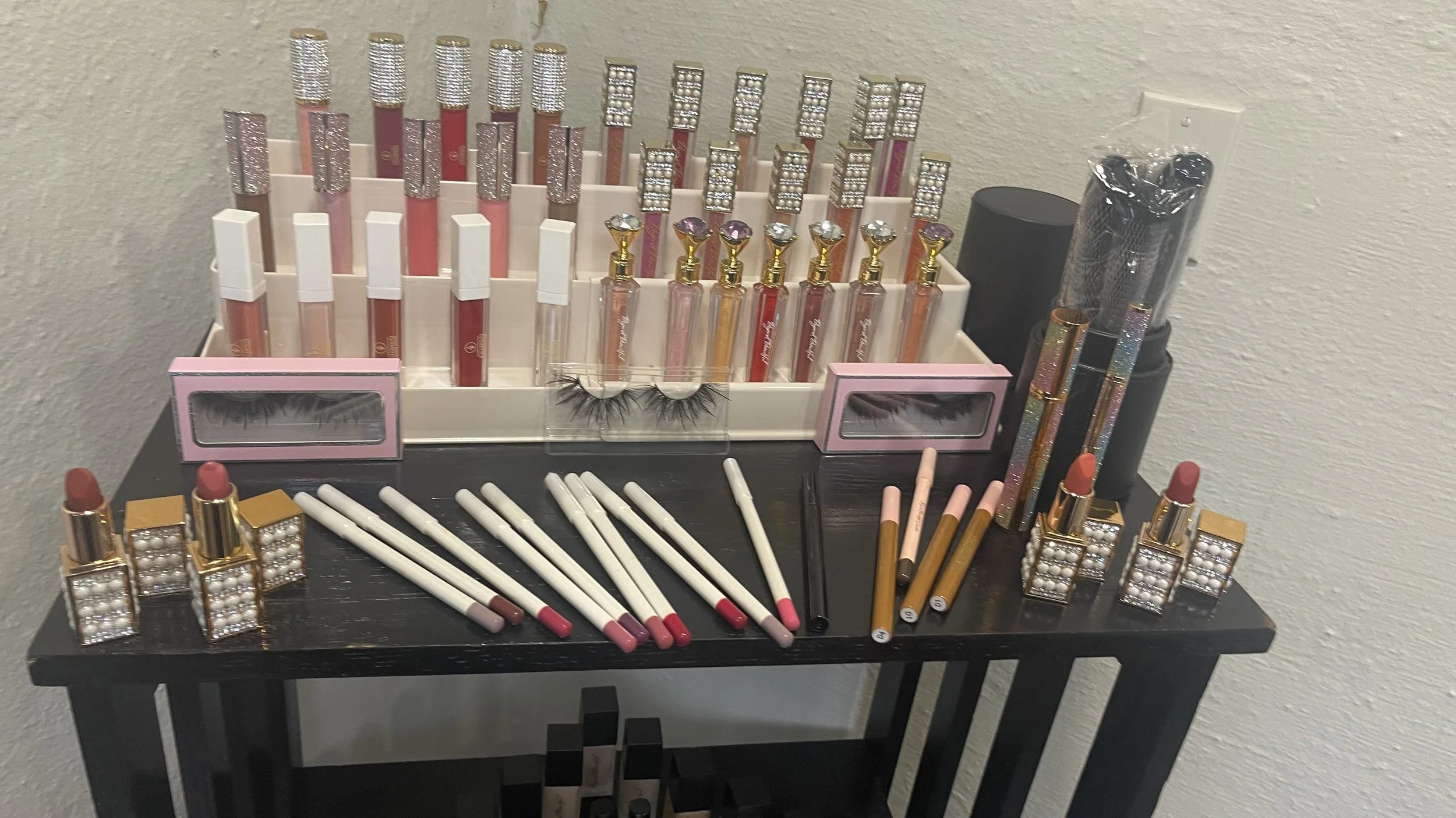 A collection of makeup and beauty products arranged on a black table with a neutral wall background. Contains lip gloss, false eyelashes, lipsticks, and various eyeliner pencils with decorative packaging.