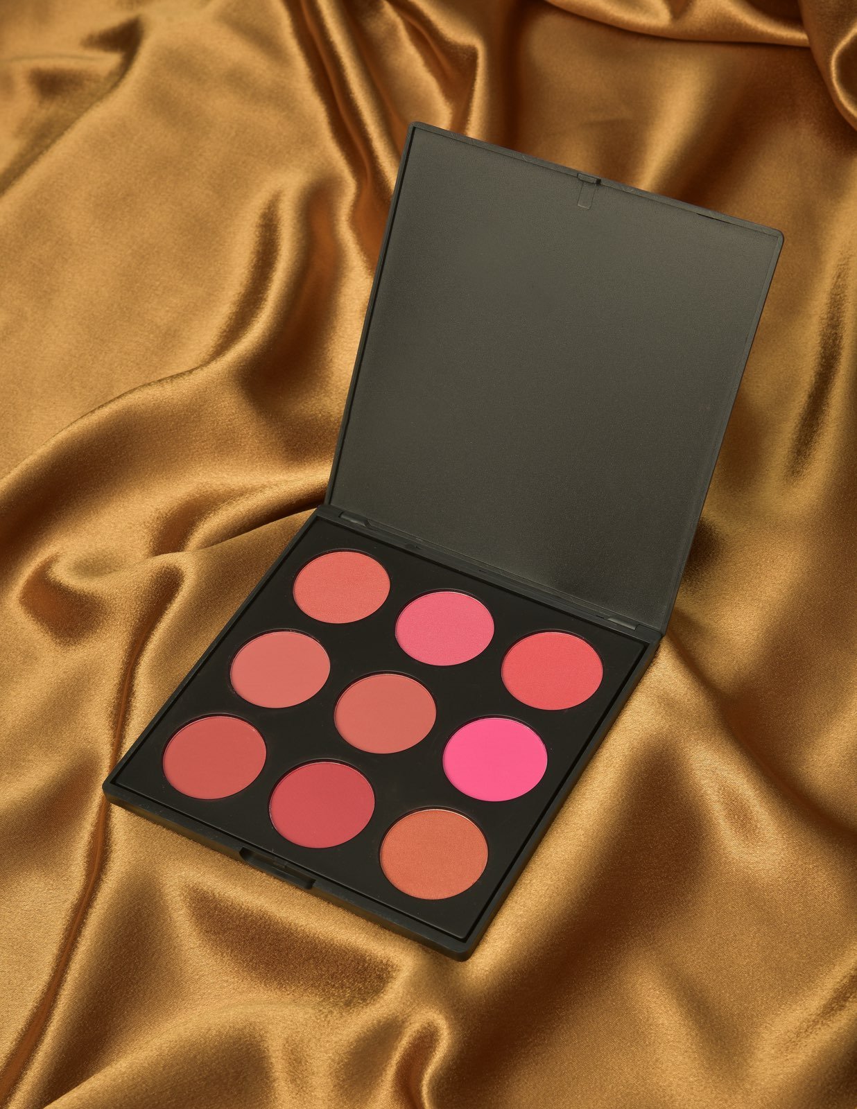 Open makeup palette with nine blush or eyeshadow shades, placed on gold satin fabric.