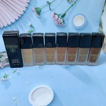 A row of seven foundations in black and transparent bottles on a blue surface, surrounded by pink flowers, a white candle, and pink paper decorations. Beyond Beautiful Cosmetics Gifts and Interactive Engagements 