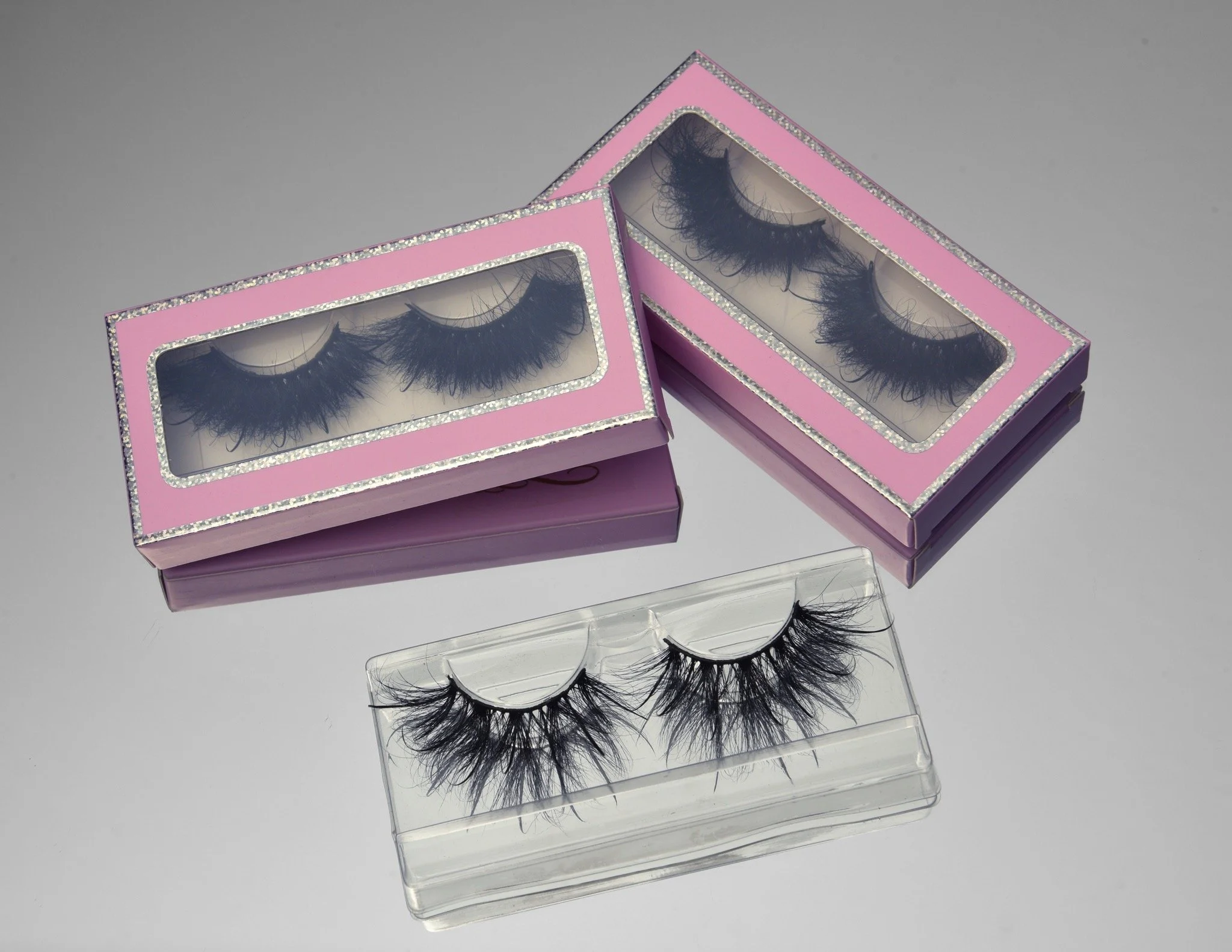 Two pink boxes with decorative edges holding black false eyelashes. Additionally, there is a plastic case containing more false eyelashes placed on a reflective surface. Eyelashes are now available at beyond beautiful.
