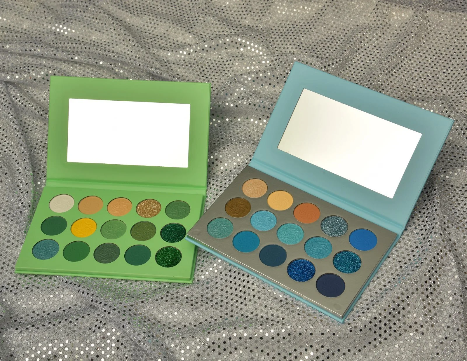 Two colorful eyeshadow palettes, one green and one blue, open to display bright and glittery shades inside, placed on a sparkly silver fabric background.