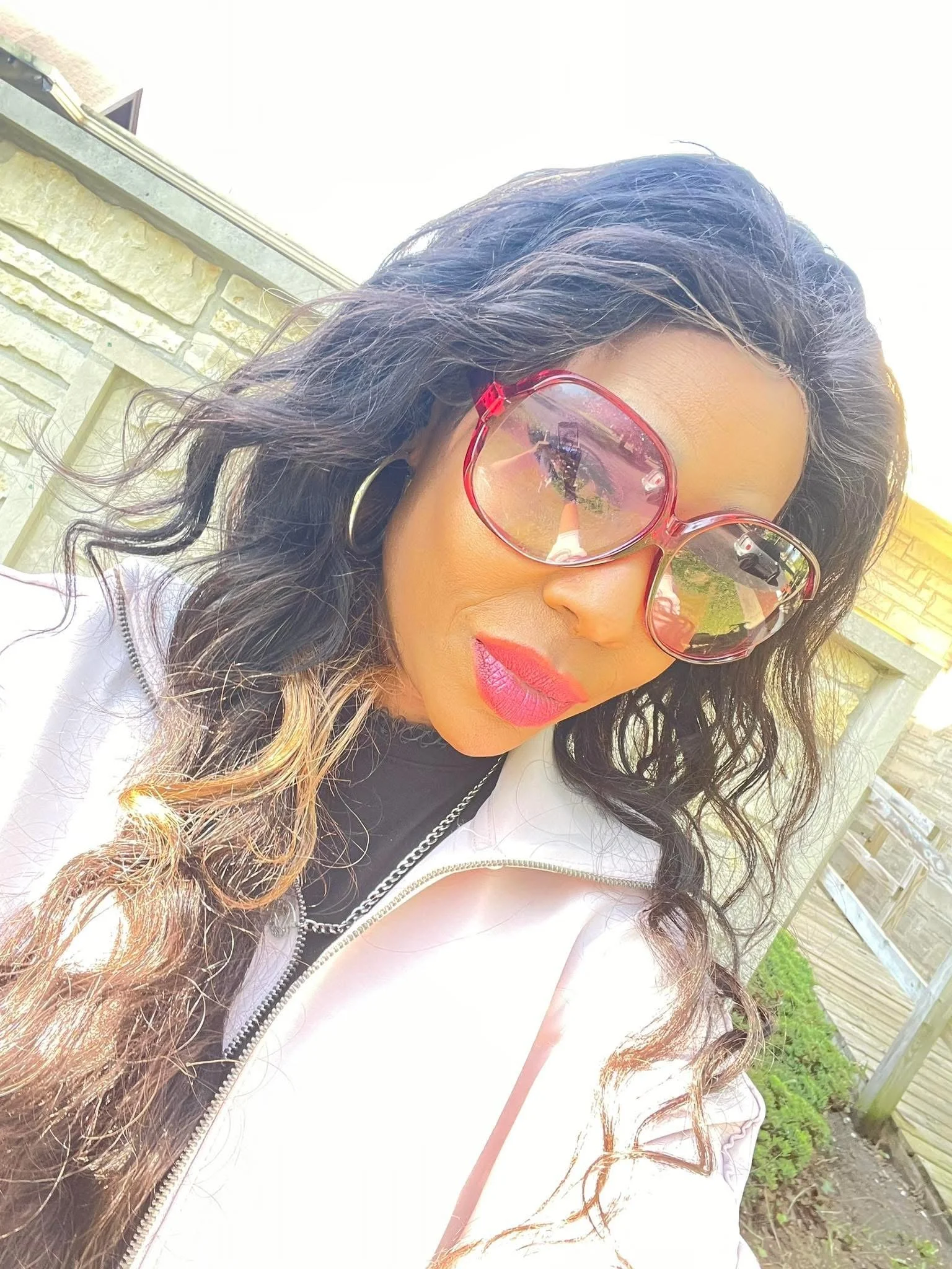 Woman with curly dark hair wearing large red sunglasses and bright pink lipstick, outdoors on a sunny day.