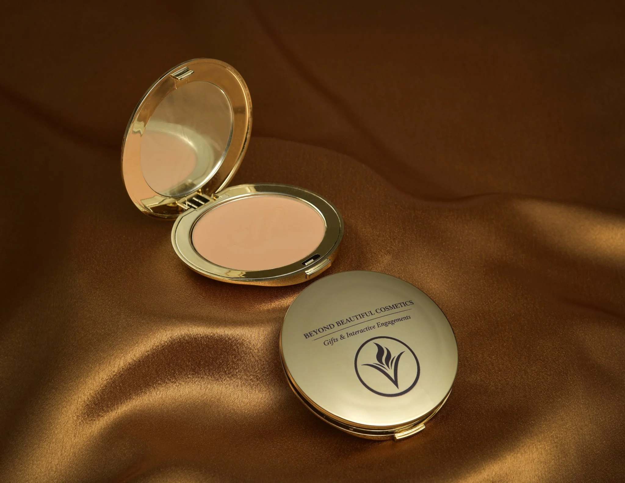 Open compact mirror on a brown fabric surface, with a round labeled sticker nearby reading "Beyond Beautiful Cosmetics."