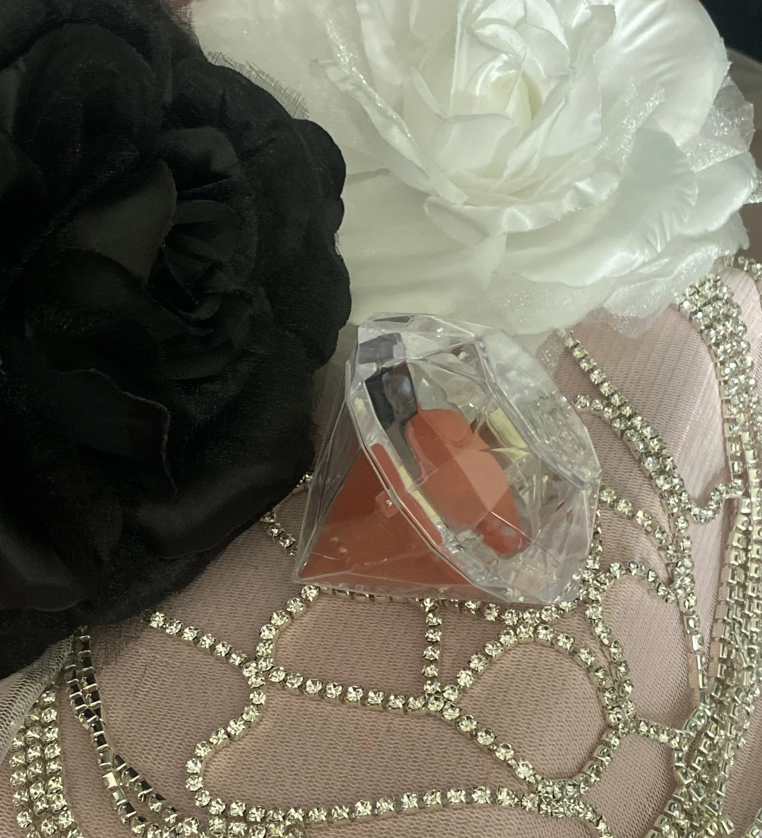 A close-up of a decorative pink surface with rhinestone accents, a white satin flower, a white satin flower with shimmering edges, a small box with a peach-colored item inside, and a gloved hand. Diamond make up sponge with case.