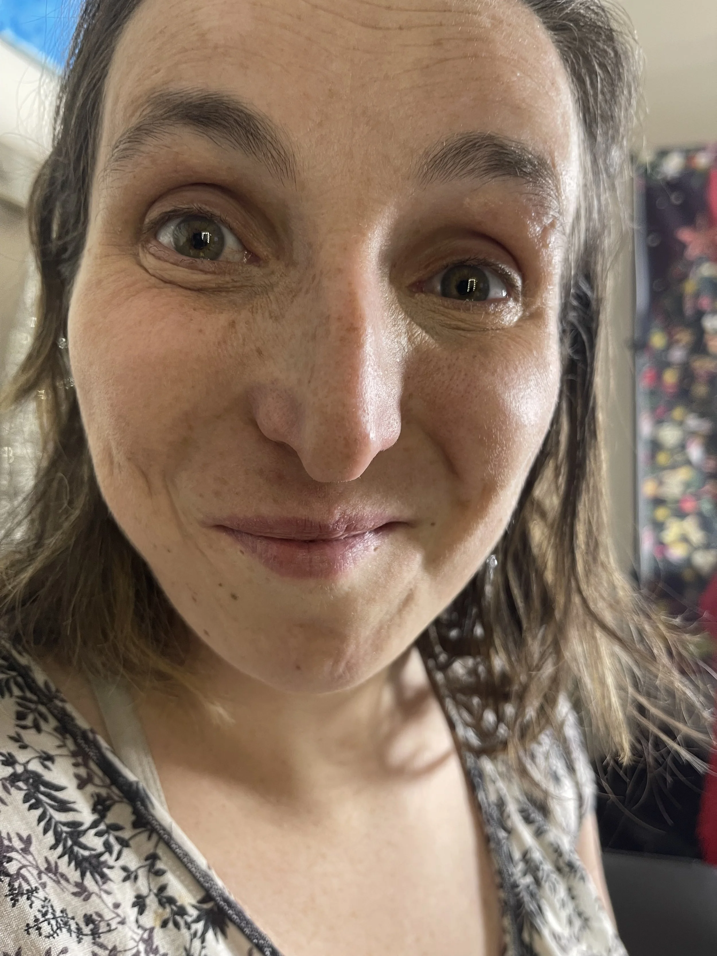 Close-up of a woman's face with light skin, brown hair, and gray eyes, smiling slightly, wearing a white and black patterned top. Customers who use Beyond Beautiful Cosmetics