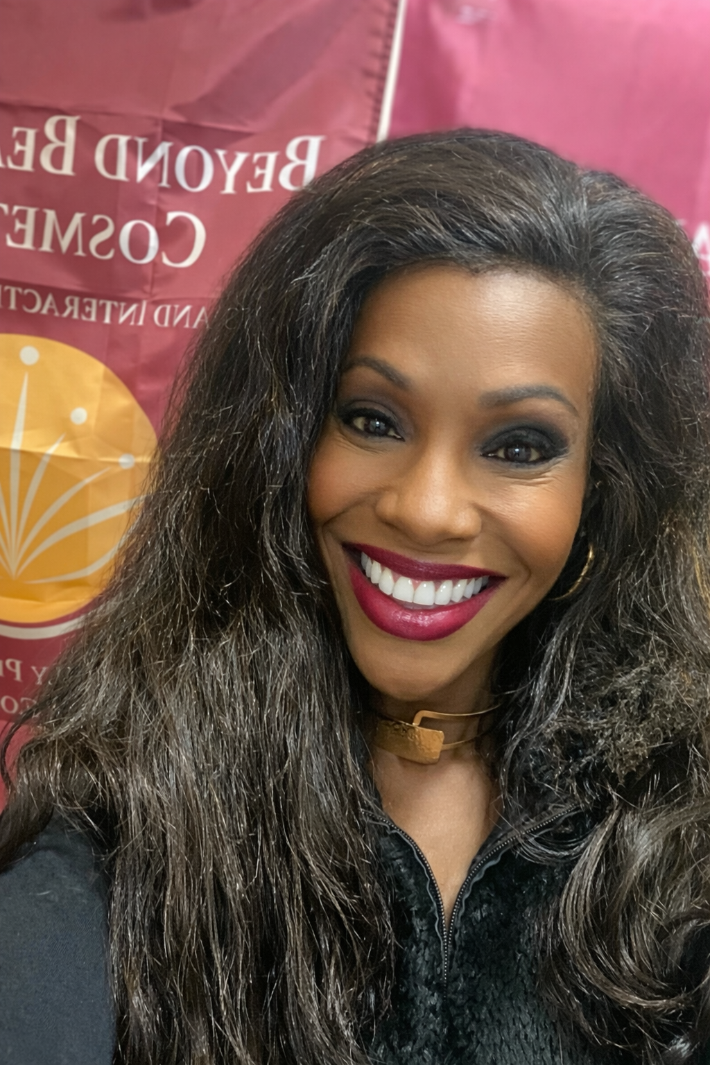 A smiling woman with long curly hair wearing a black outfit and gold jewelry, standing in front of a maroon and yellow banner. Beyond Beautiful Cosmetics Gifts and Interactive Engagements Founder Baiyina Hodge