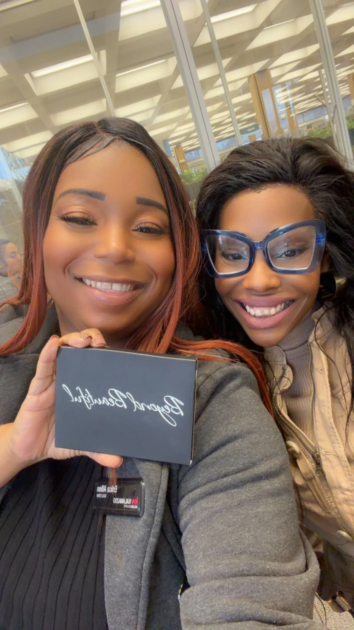 Two women sitting close together, smiling at the camera, one holding a black box with white cursive writing, in an indoor setting with glass walls and ceiling panels. Customers who use Beyond Beautiful Cosmetics