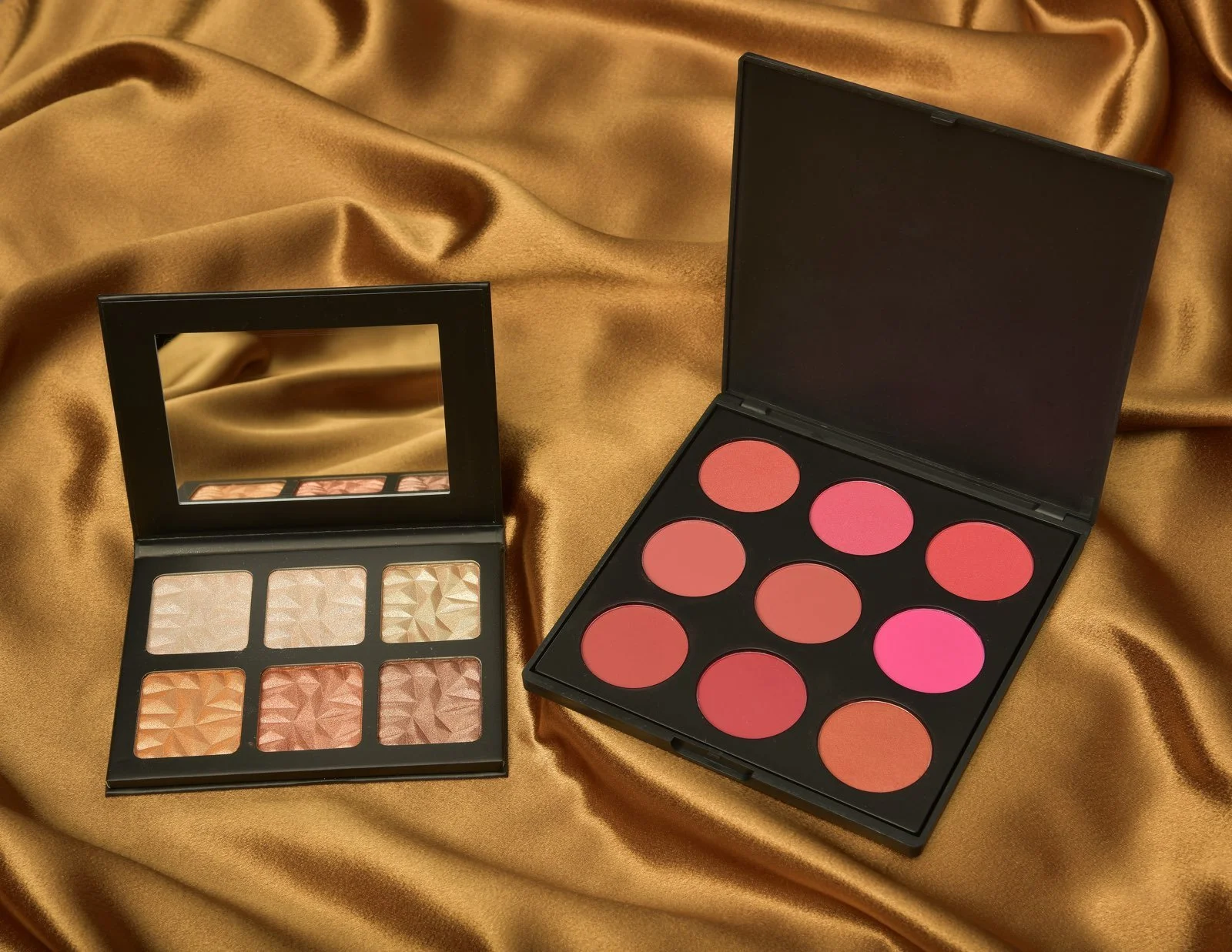 Two makeup palettes on a satin gold-colored fabric. One palette contains neutral and shimmery eyeshadows, and the other contains pink and red blushes.