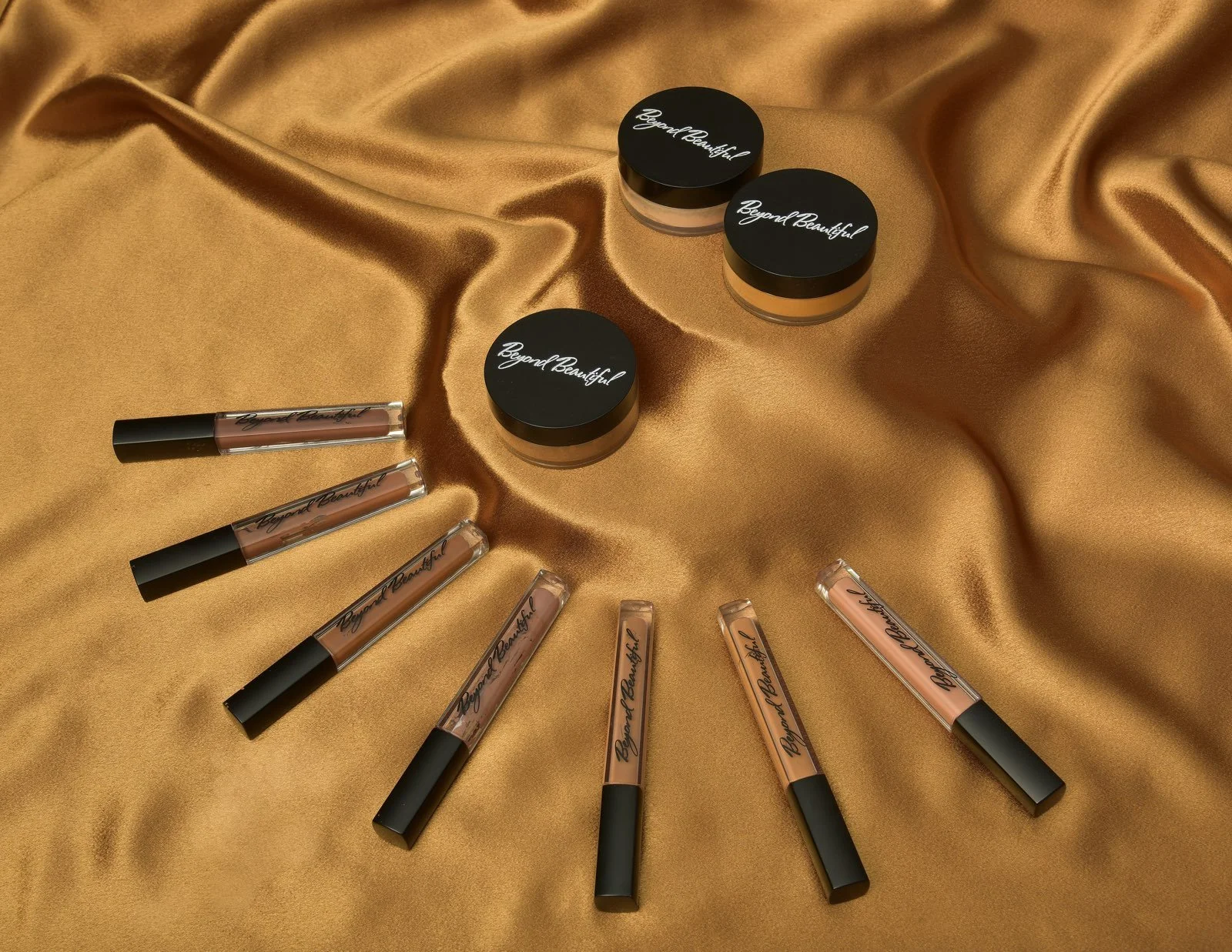 A collection of makeup products, including multiple lip glosses and face powders, placed on a gold satin fabric. The lip gloss tubes are arranged in a semi-circle, and the face powders are in small round containers with black lids labeled 'Beyond Bea