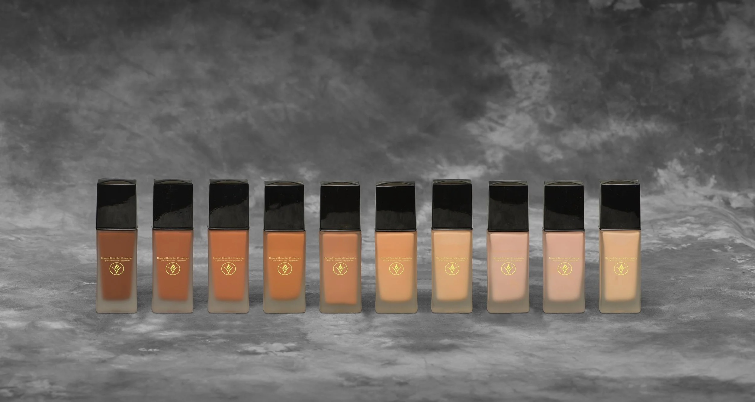 A line of ten foundation bottles with black caps, varying shades from dark to light, placed on a gray marble surface against a gray cloudy background.