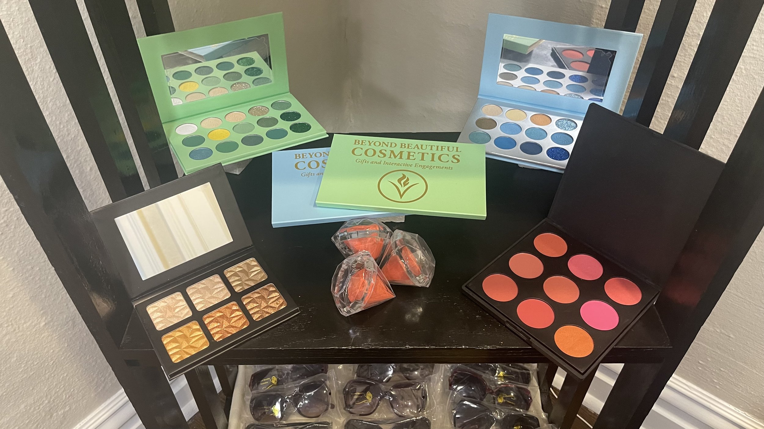 Display of various makeup palettes and cosmetics on a black wooden shelf, including eyeshadows in green, blue, orange, and gold shades, with some in clear plastic packaging.