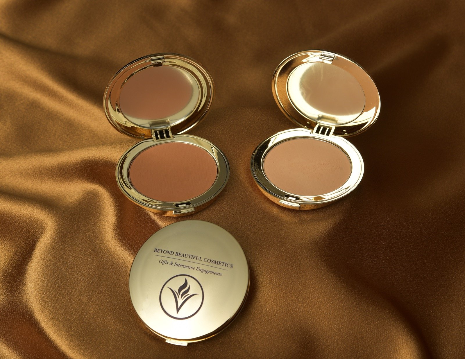 Two mirrored compact cases on a satin surface and a white lid with black text that reads "BEYOND BEAUTIFUL COSMETICS" and a logo.