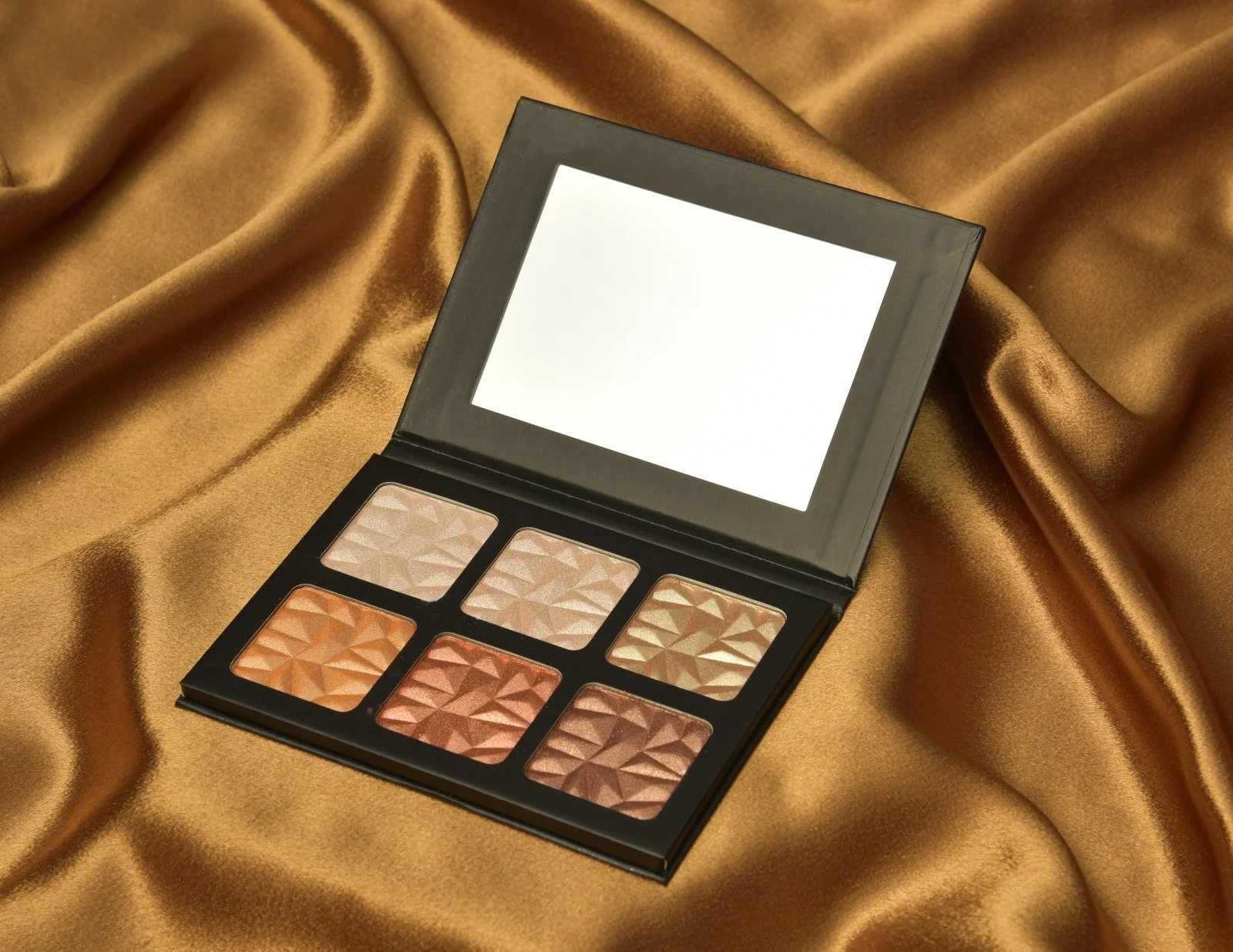 An open makeup palette with six shades of eyeshadow, placed on a gold satin fabric.