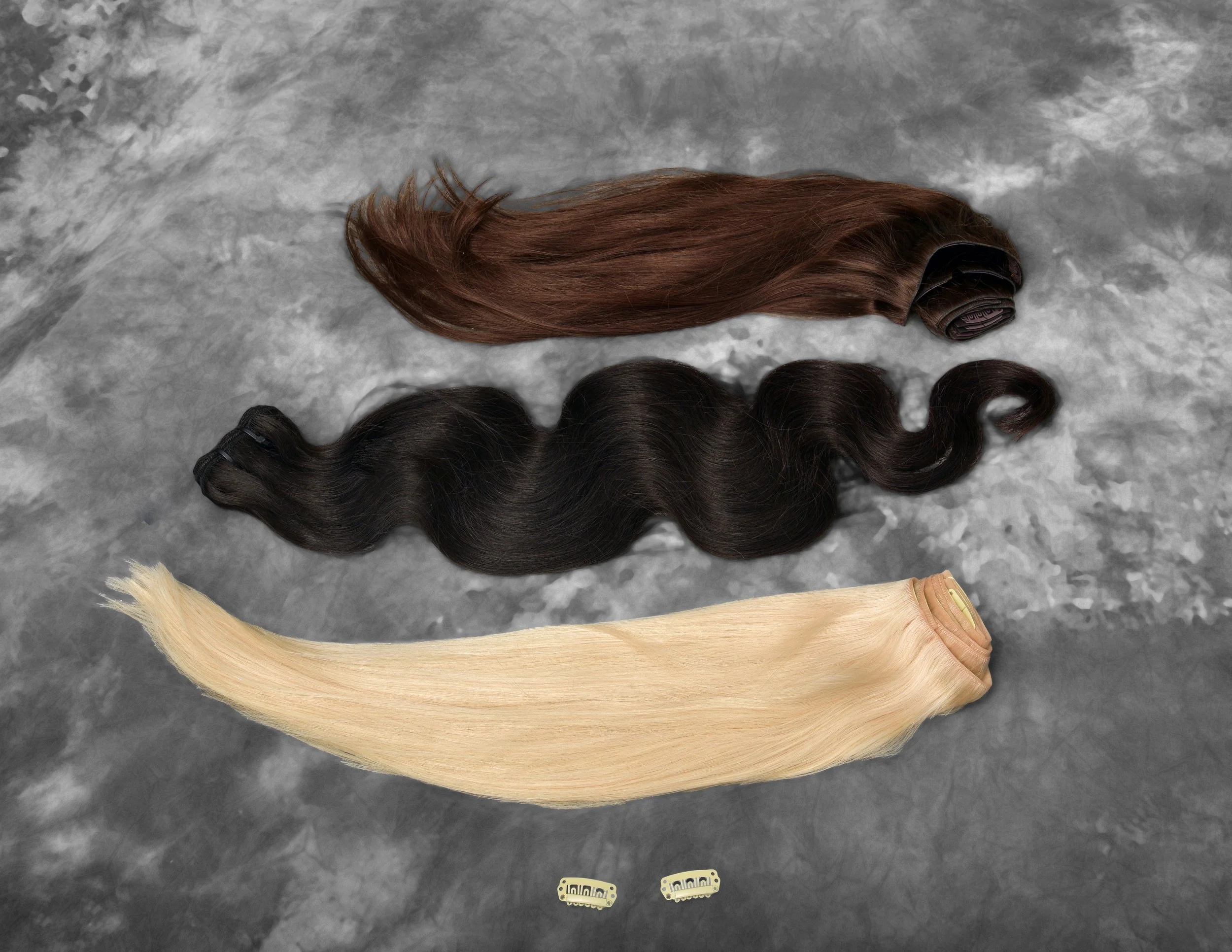 Four bundles of hair extensions in various colors, placed on a gray surface. The colors include black, brown, light brown, and blonde. Hair bundles and clippings are now available at beyond beautiful.