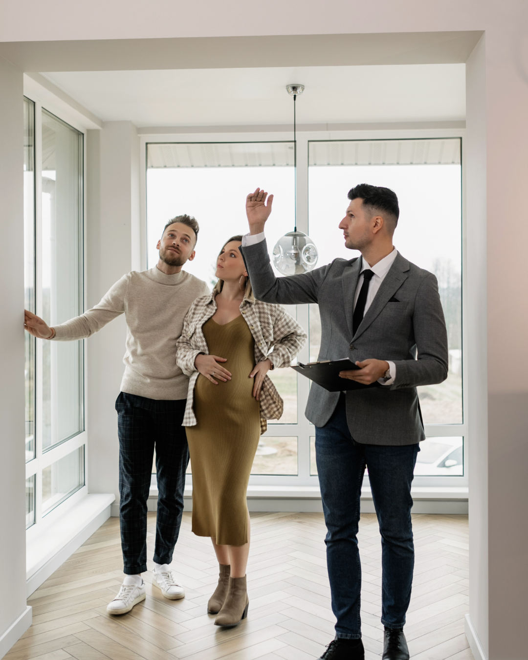 A couple is talking to a real estate agent in a new house. The man is wearing a beige sweater and plaid pants, the woman is pregnant and wearing a green dress with a plaid shirt, and the agent is dressed in a gray suit with a clipboard, raising his h