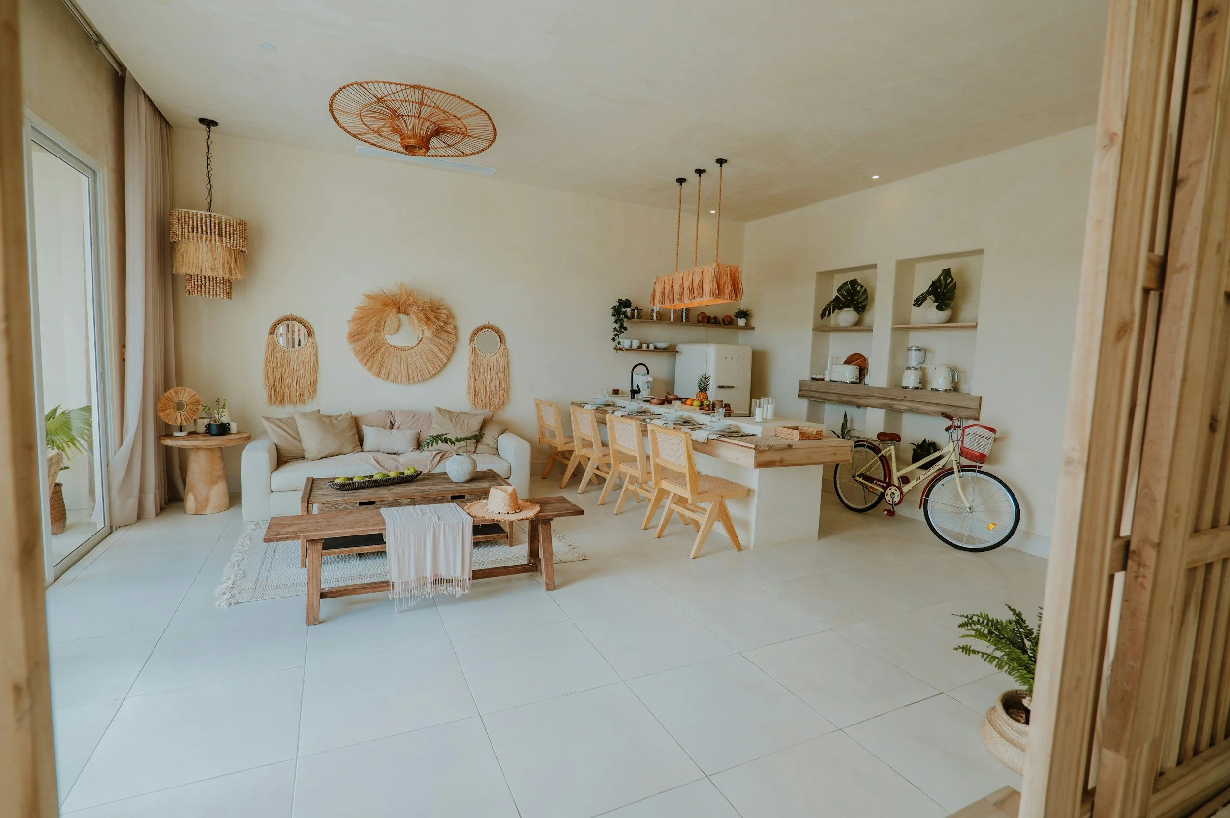 Bright, modern living room with neutral tones, a white sofa, wooden coffee table, wall decor with natural woven art, and a dining area with wooden chairs and a white kitchen with open shelves and plants.