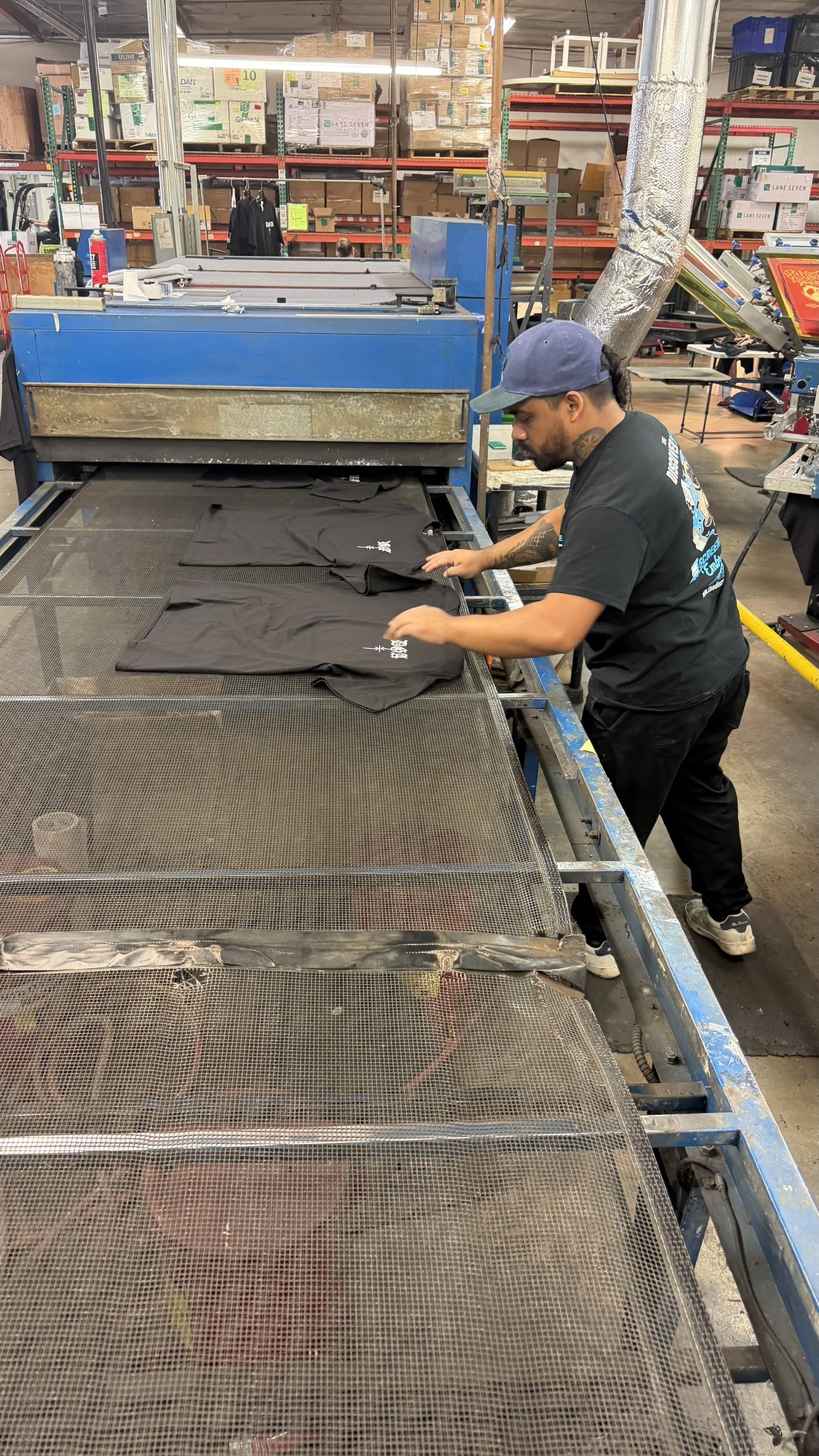 A man working on a T-shirt printing machine in a warehouse or print shop, with stacks of boxes and shelves in the background.