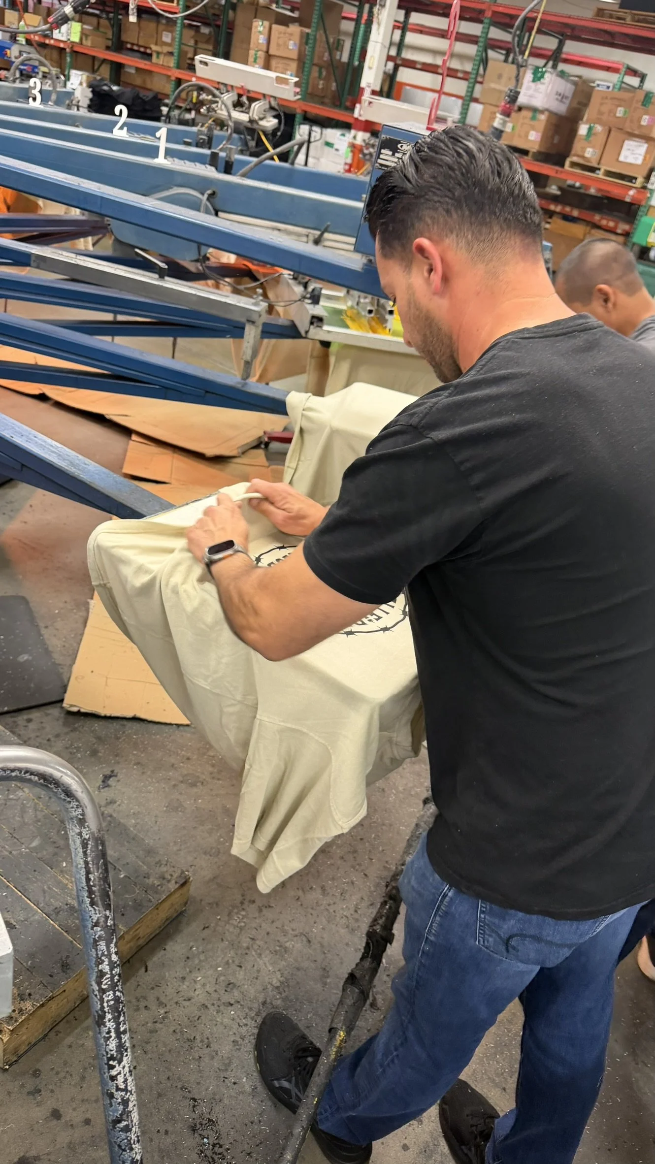 A man in a black shirt and blue jeans is folding a beige T-shirt with a printed logo in a warehouse or printing shop. There are blue printing presses and shelves with boxes in the background.