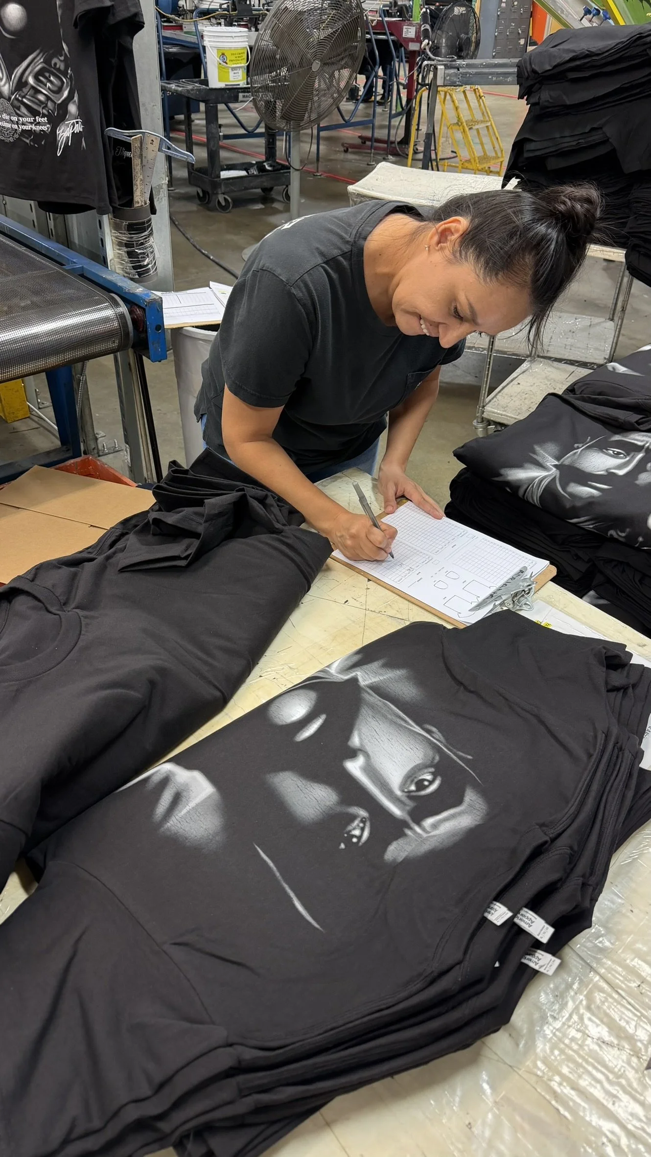 A woman working in a t-shirt production facility, signing a clipboard surrounded by neatly folded black t-shirts with artwork, with industrial equipment and tools in the background.