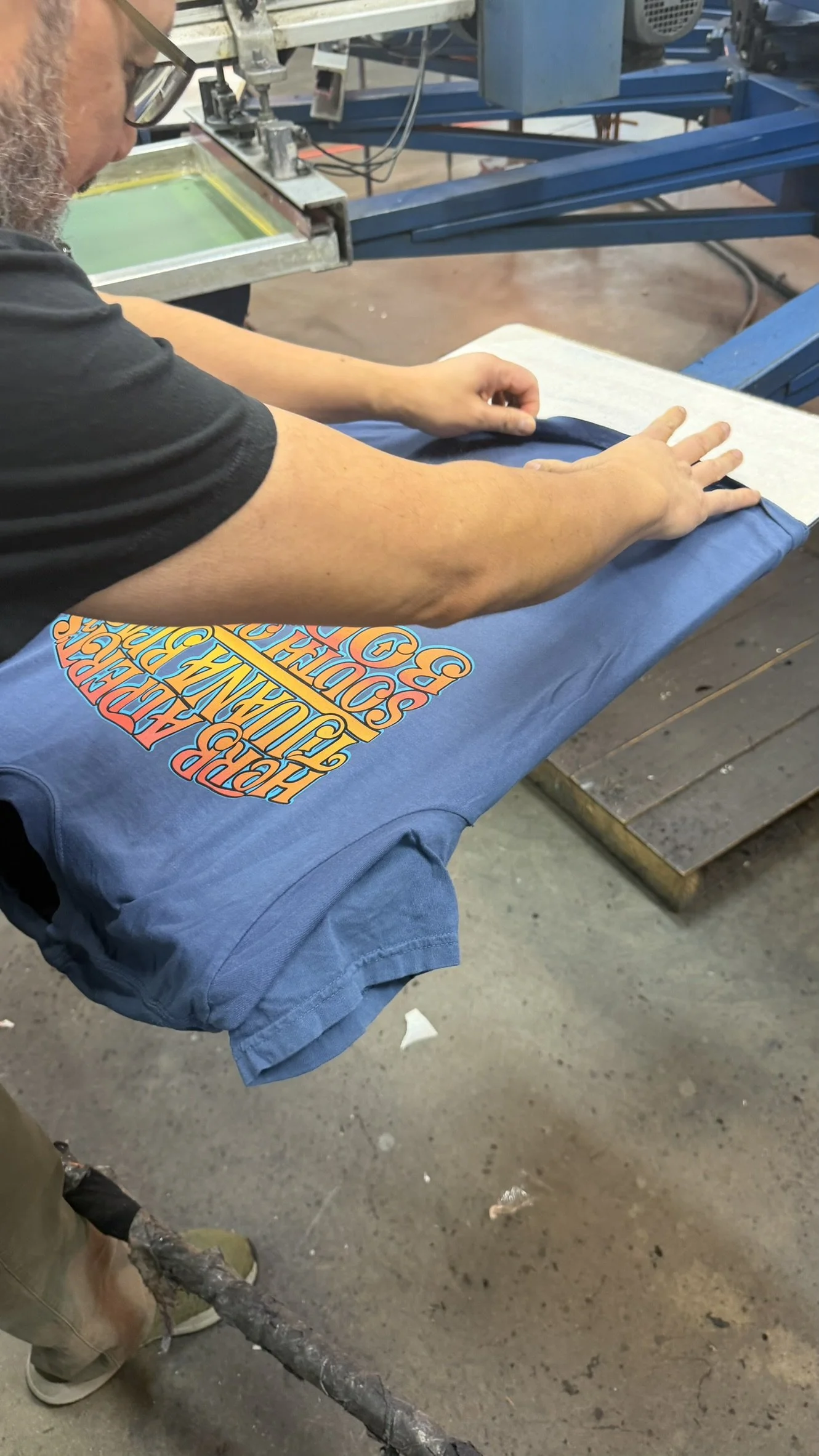 Person folding a blue T-shirt with a colorful graphic design on it at a printing or manufacturing facility.