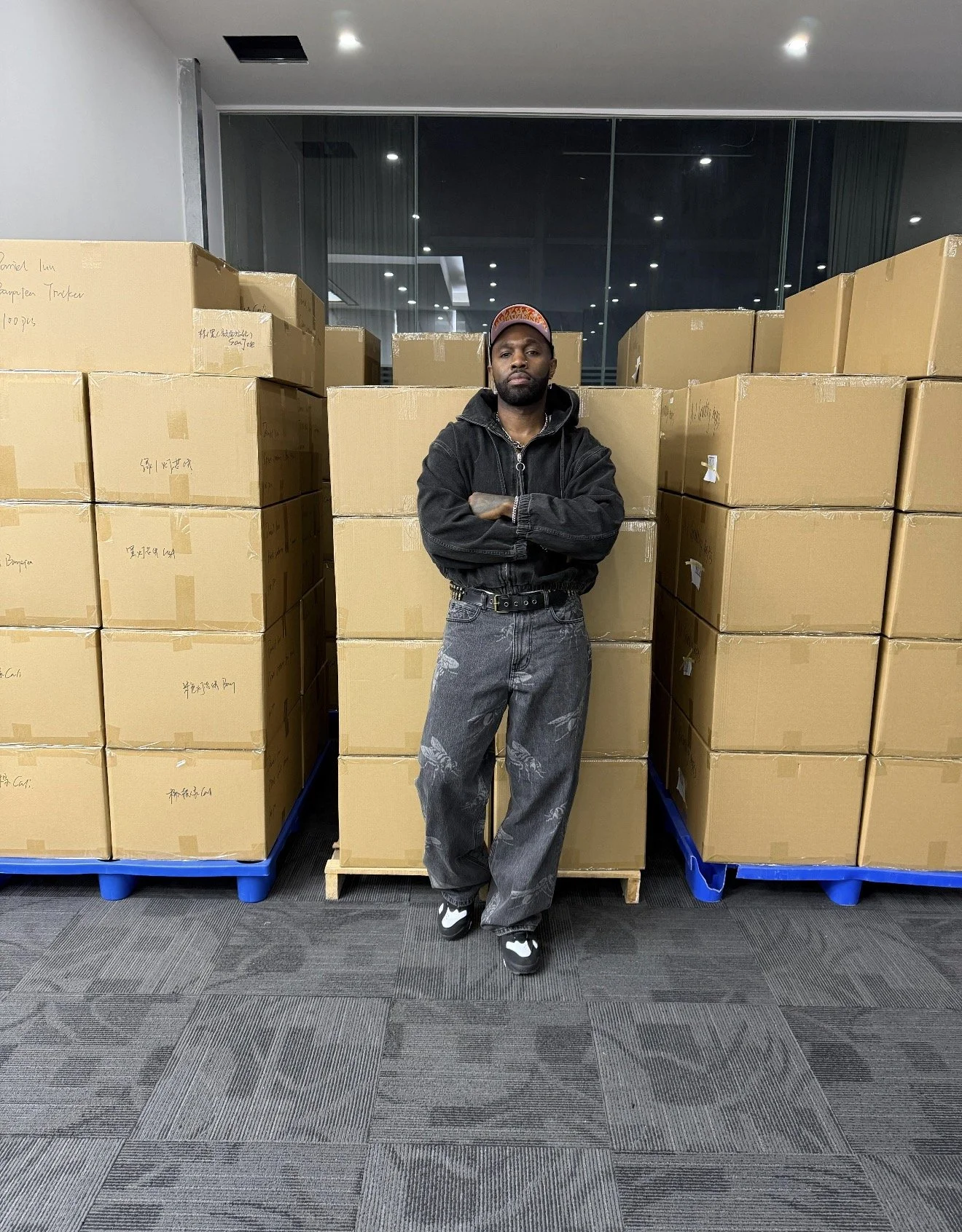 A man stands with arms crossed in front of stacked cardboard boxes in a warehouse or storage room, with a large glass window and ceiling lights in the background.