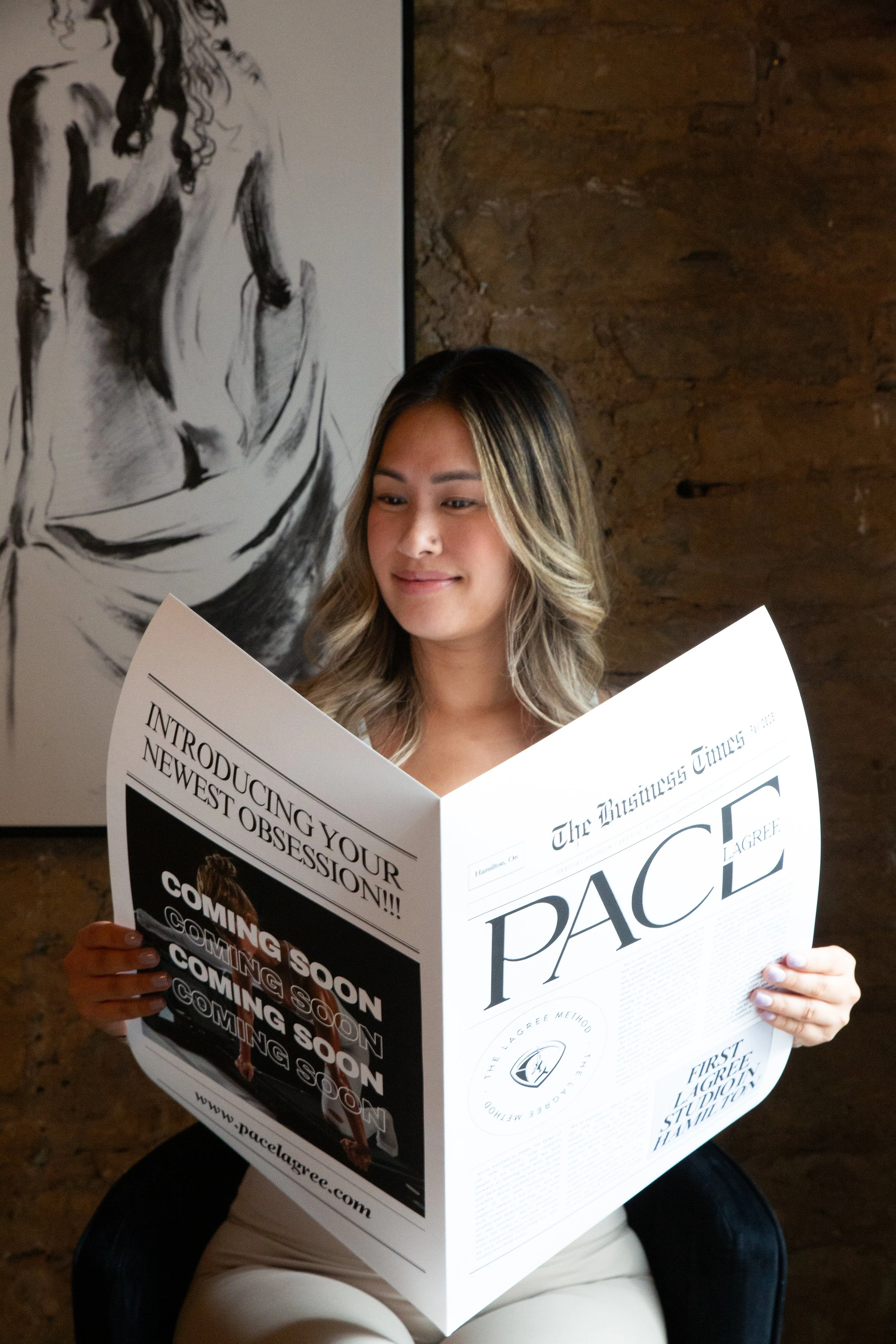 A woman with shoulder-length blonde hair and light skin reading a newspaper titled 'The Pacific Times' in a room with a brick wall and a black-and-white artwork of a woman.