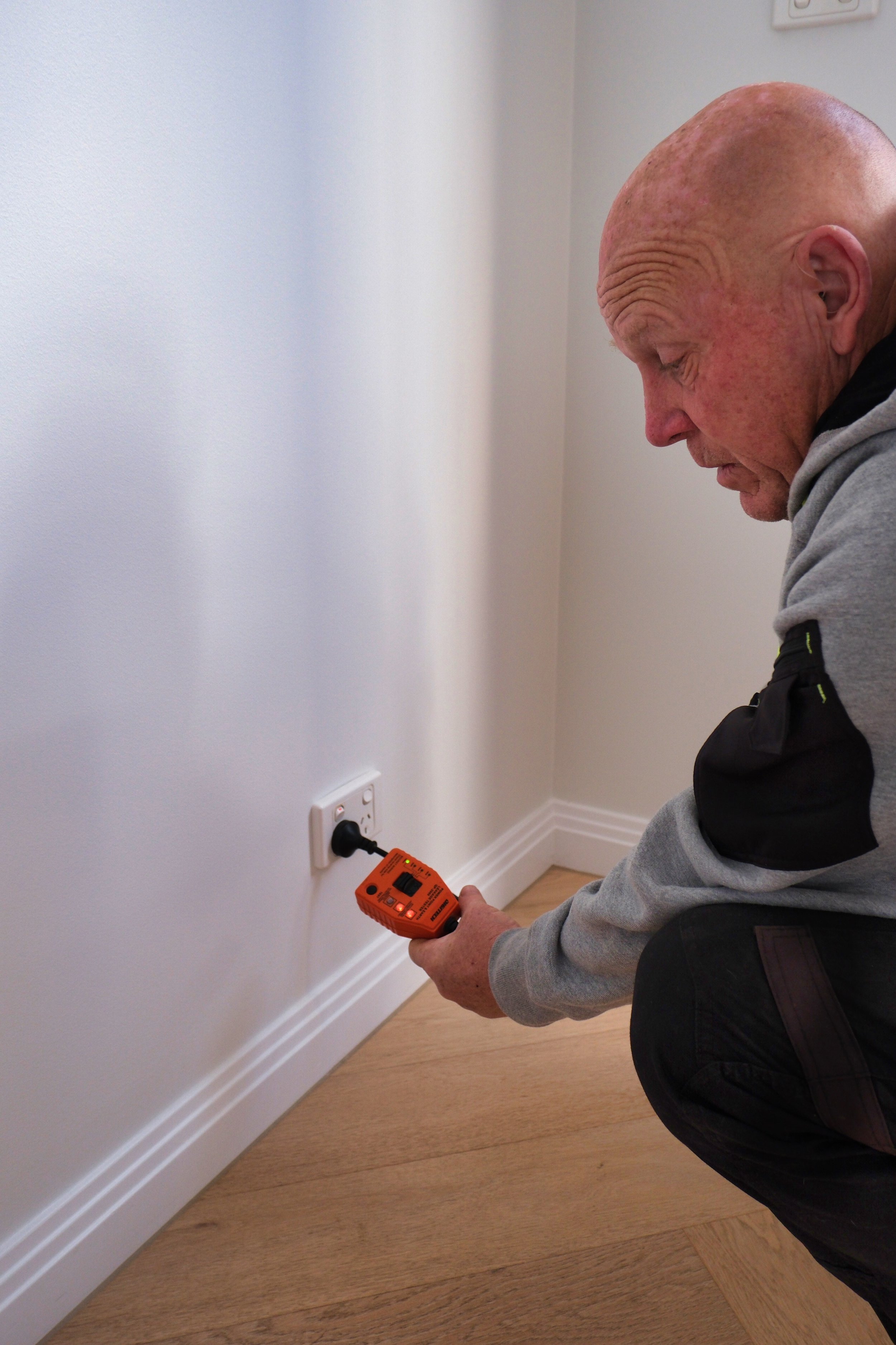 A man crouches down using a voltage tester to check an electrical outlet in a room with white walls and wooden flooring.