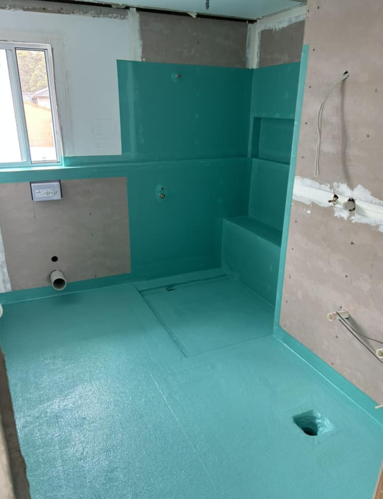 Interior of a bathroom under construction with turquoise waterproofing on the walls and floor, plumbing pipes visible, and a window on the left side.