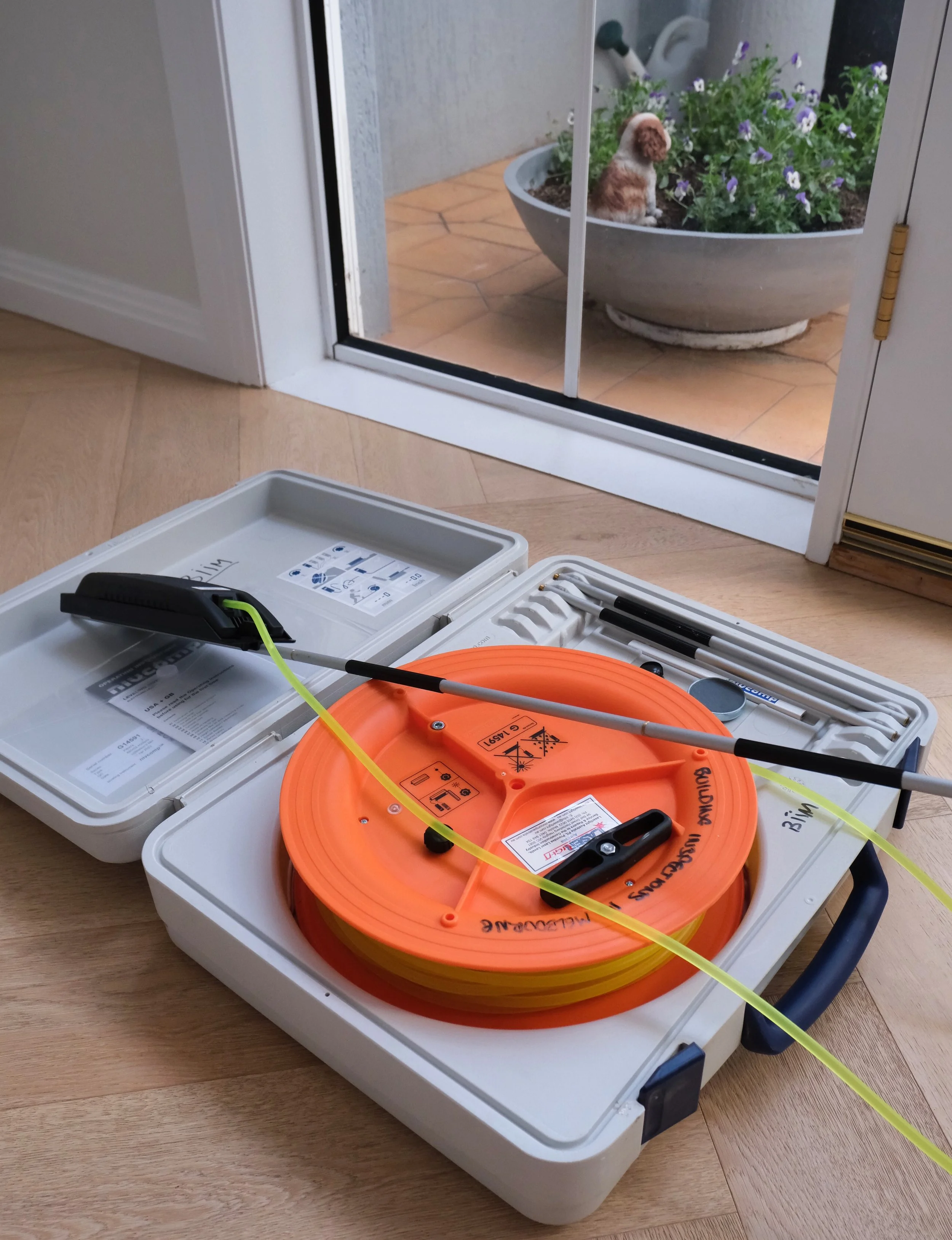 A black and yellow drain snake tool with a long flexible metal cable inside an orange reel, placed on a white carrying case on a wooden floor. The case is open, revealing compartments and instruction sheets. In the background, a glass sliding door sh