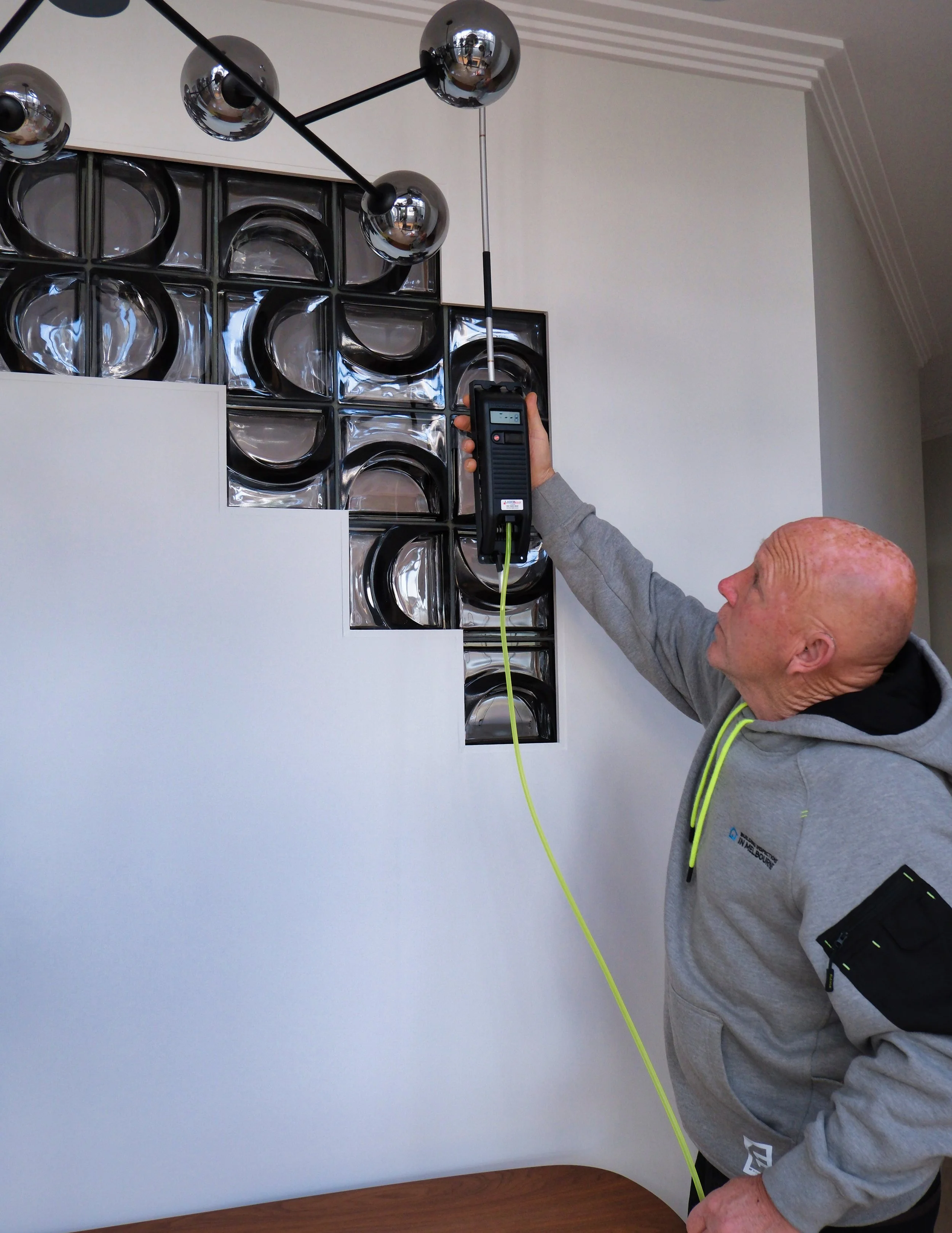 A man in a gray hoodie is holding a testing device with a yellow cord, testing a modern black and chrome pendant light fixture mounted on the ceiling of a room with white walls and decorative molding.