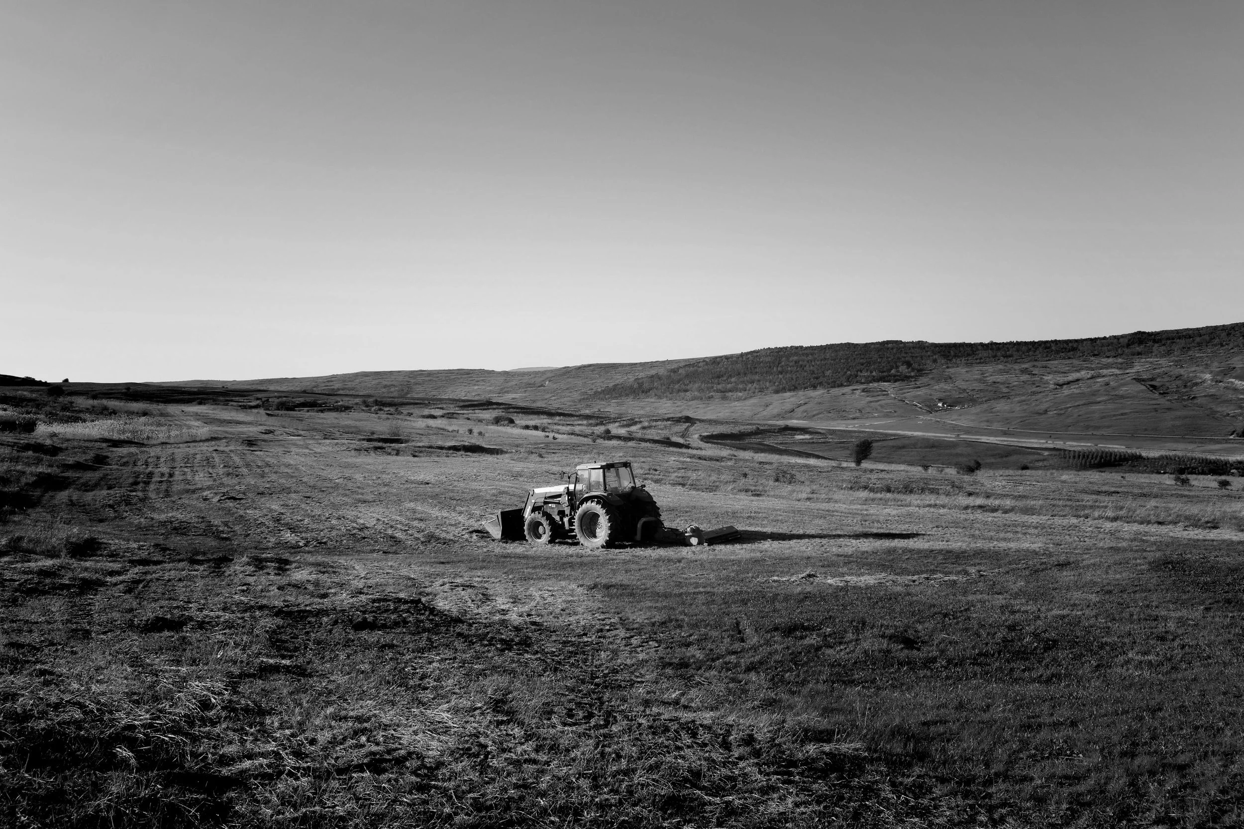 A tractor working in an open field during daytime, with rolling hills in the background, depicted in black and white.