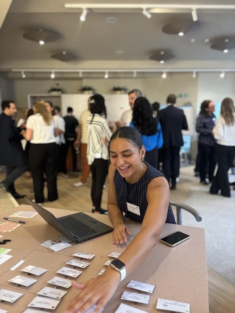 Young woman smiling and reaching out to pick up a name badge at a professional networking event or conference, with other attendees in the background.