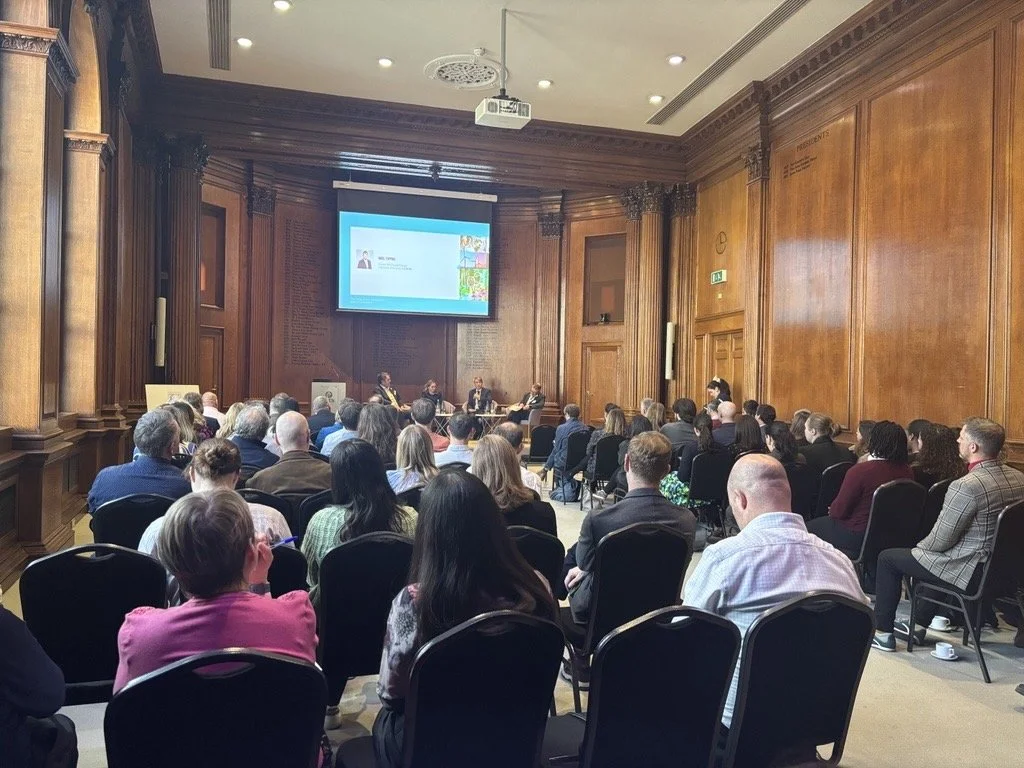 A conference room with wooden panel walls filled with an audience attending a presentation. There is a large screen displaying a slide at the front of the room.
