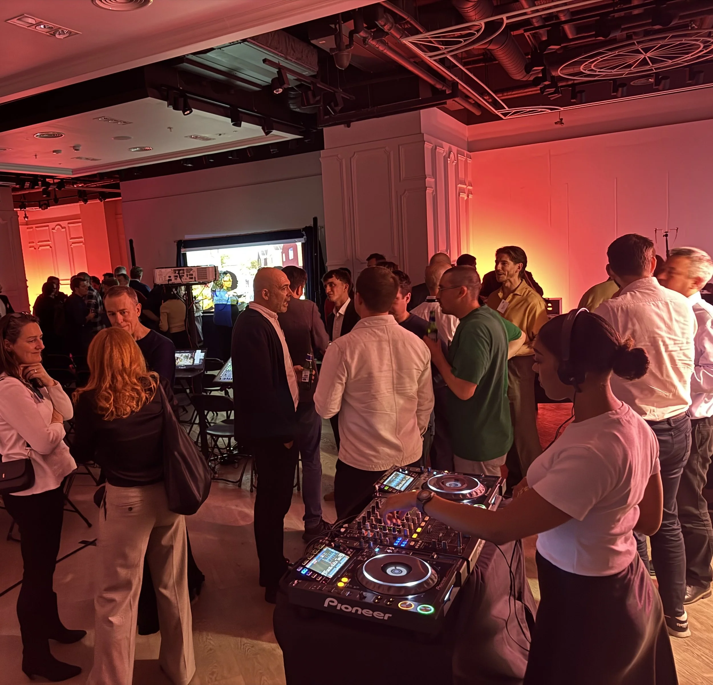 People socializing at a party or event, with a DJ playing music on a Pioneer DJ controller in the foreground, colorful lighting, and a projected screen in the background.