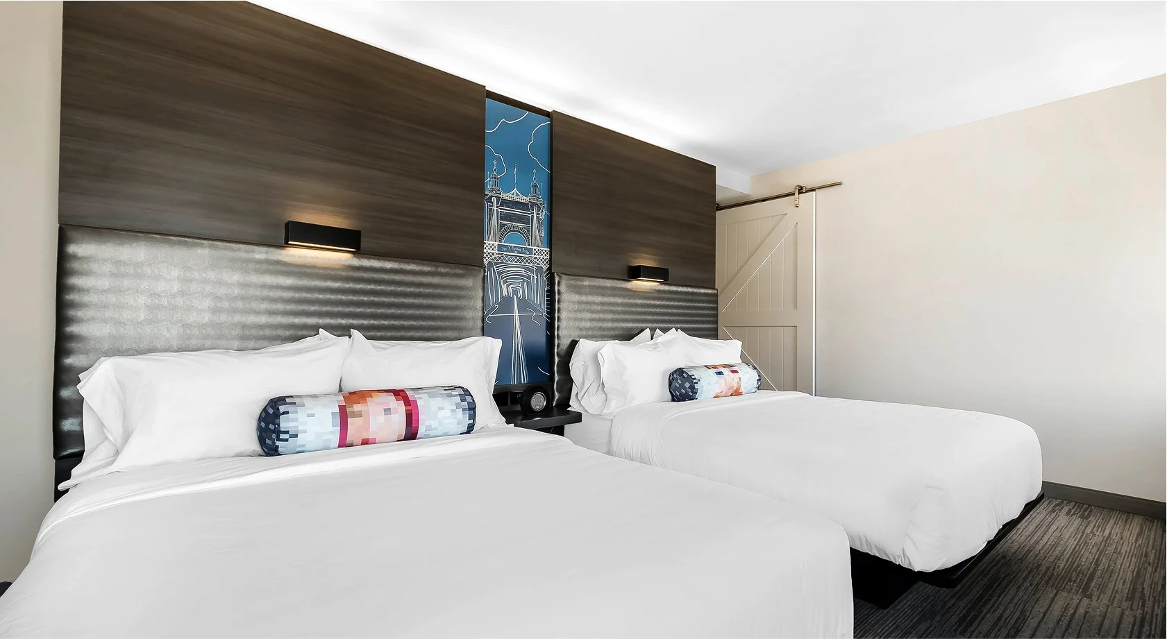 Two neatly made beds with white bedding, decorative pillows, and a large padded headboard, in a modern hotel room featuring a wood-paneled wall, wall-mounted lights, and a slide door.