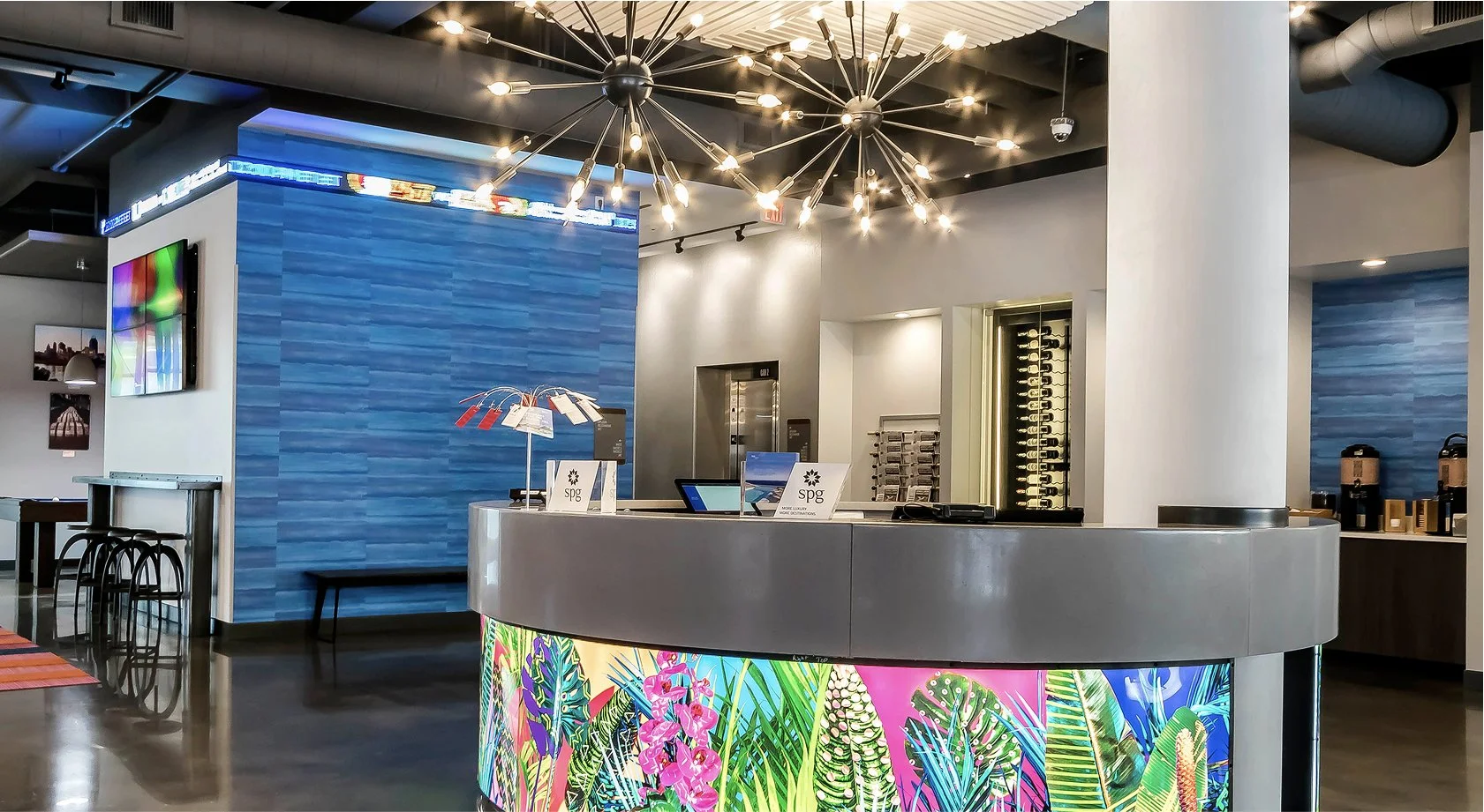 A modern hotel lobby reception area with a curved front desk decorated with a vibrant tropical floral pattern. Behind the desk, there is a blue textured wall, a monitor, and a brochure stand. The ceiling features a unique light fixture with multiple