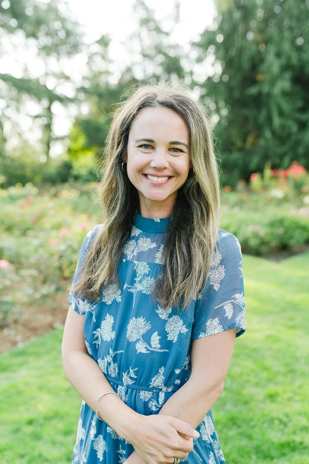 A woman with long light brown hair, smiling, standing outdoors on a grassy lawn with trees and flowers in the background, wearing a blue dress with white floral patterns.