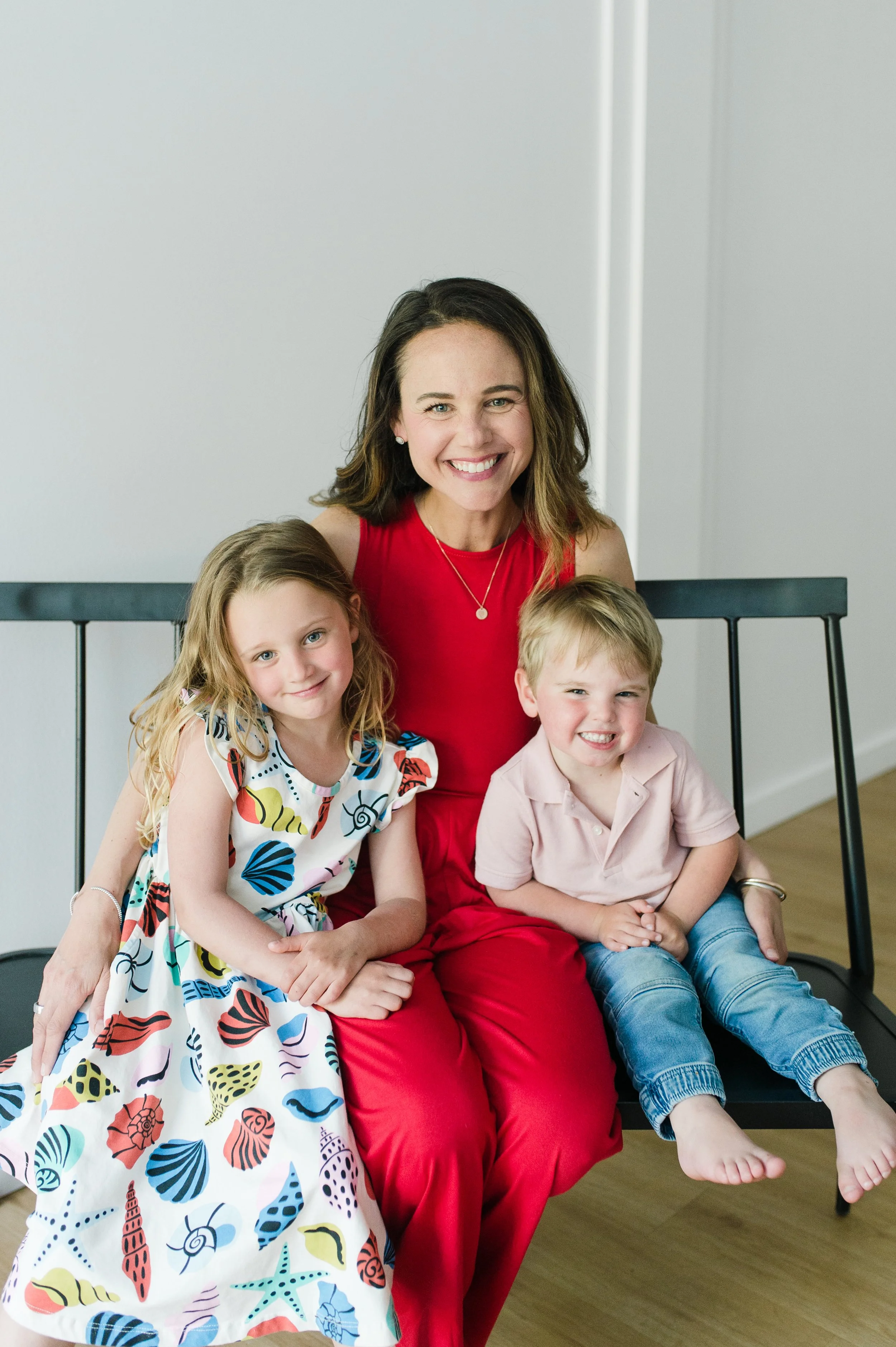 A woman in a red dress sitting on a black bench with two children, a girl and a boy, smiling at the camera in a bright indoor setting.