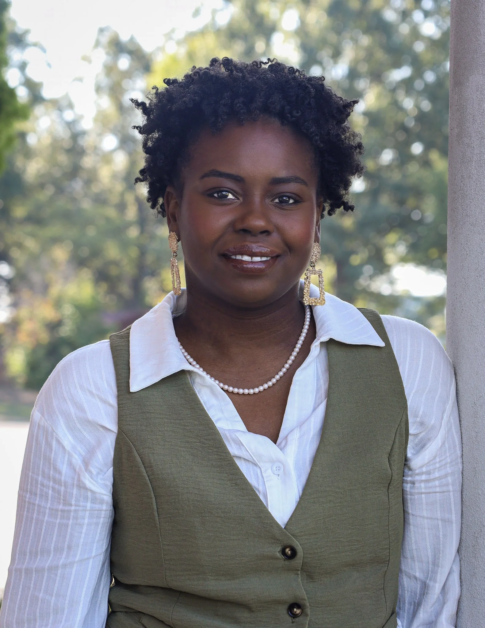 A woman with short, curly black hair smiling outdoors with green trees in the background. She wears earrings, a pearl necklace, a white shirt, and a green vest.