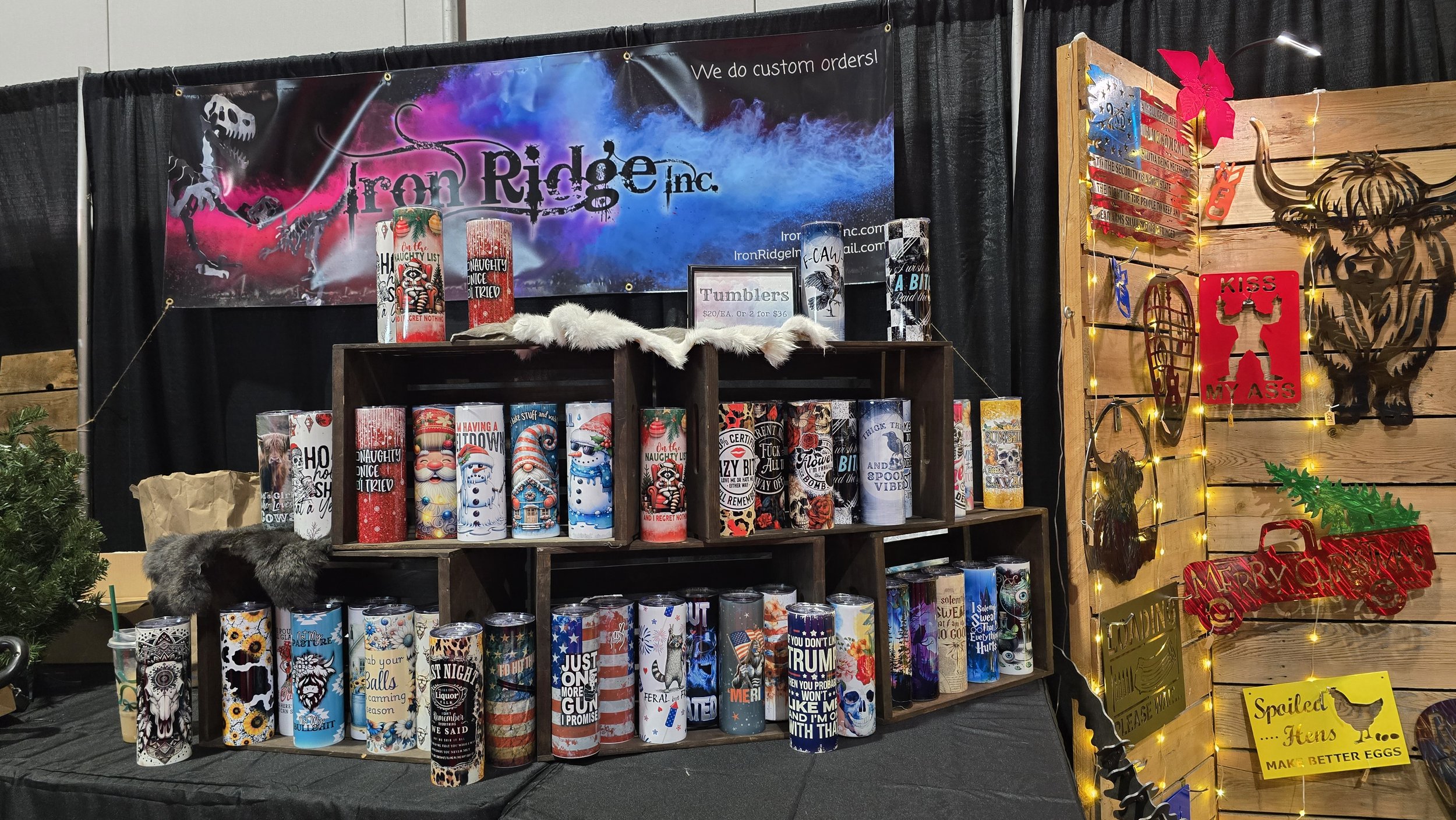Display of decorative tumblers with Christmas-themed designs at a booth, with a banner reading 'Iron Ridge Inc.' in the background and Christmas decorations on the side.
