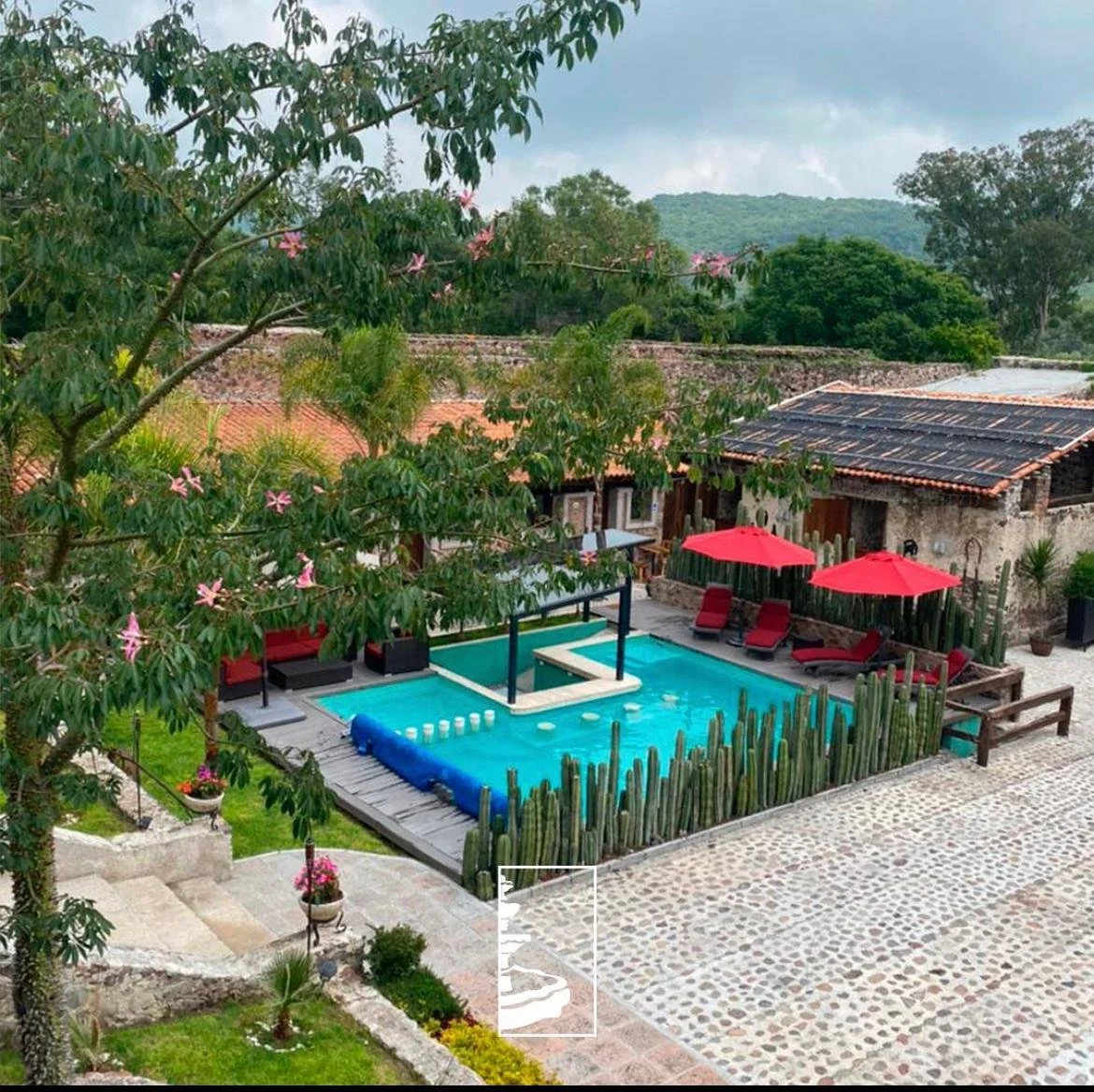 Piscina en un patio con tumbonas y sombrillas rojas, rodeada de plantas y casas con techos de teja, en un entorno rural con montañas en el fondo.
