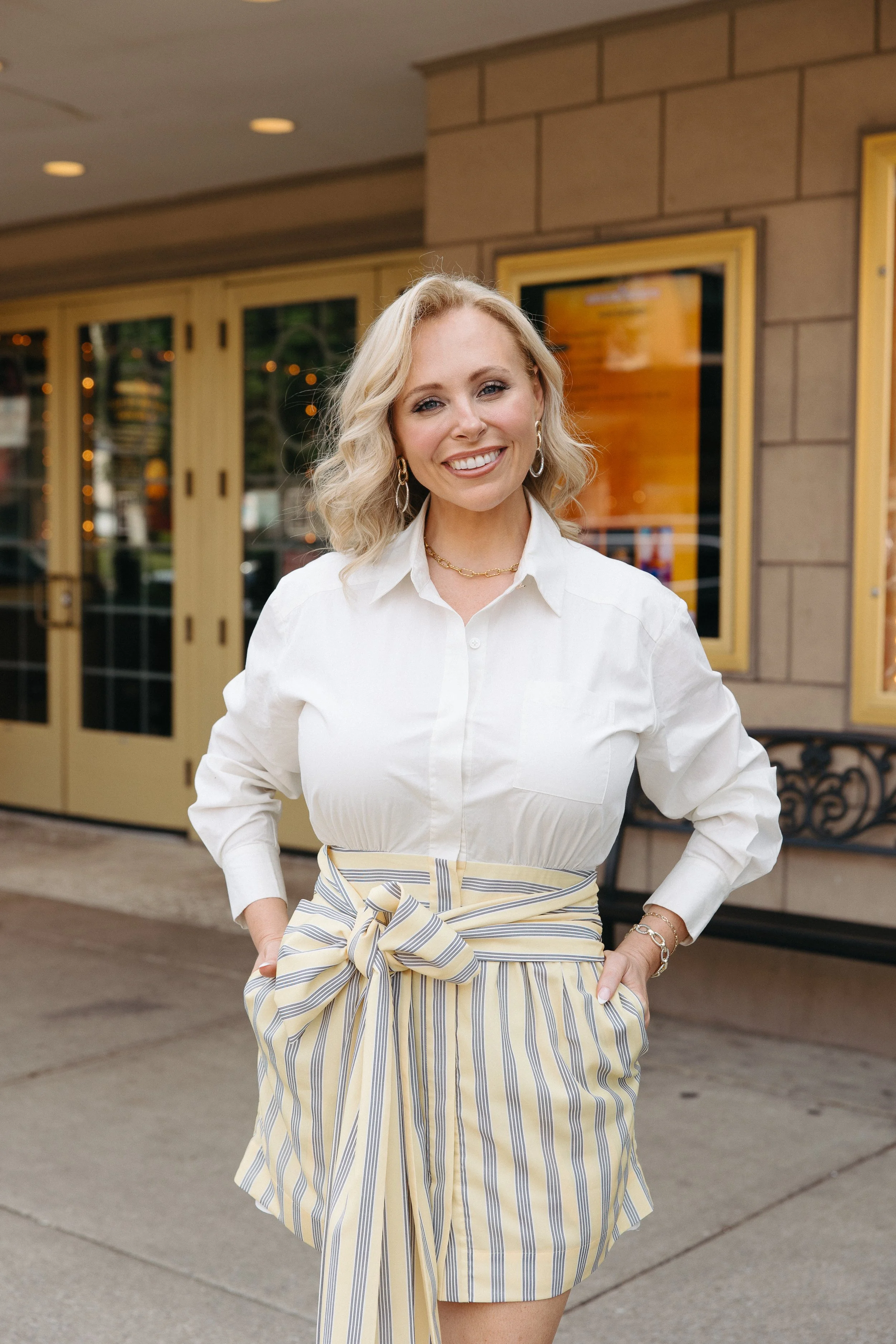 A woman with blonde, wavy hair, wearing a white shirt and striped skirt with a large bow, standing outside of a building with a glass door and framed posters.