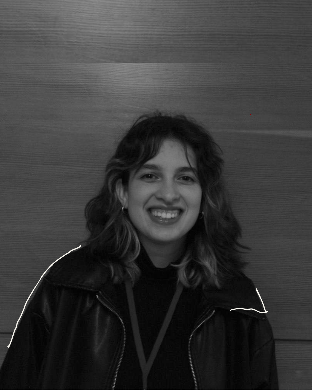 Black and white photo of a young woman with wavy hair smiling, wearing earrings, a dark jacket, and a lanyard, standing against a wooden wall background.