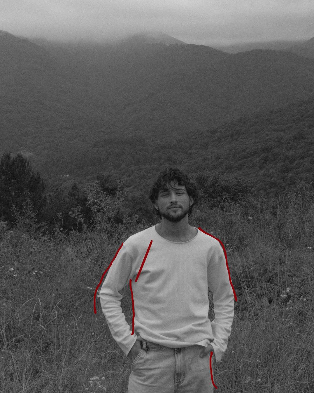A black and white photo of a young man with curly hair and a beard standing outdoors in a grassy area with mountains in the background. His hands are in his pockets and he is wearing a long-sleeved shirt and jeans.