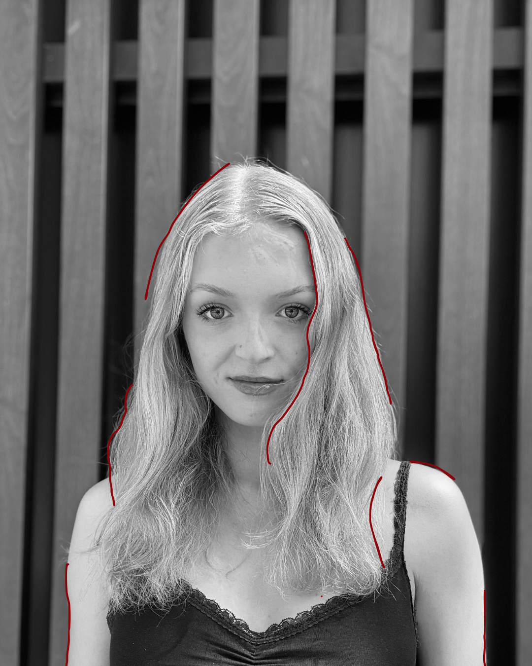 Black and white portrait of a young woman with long, wavy hair, wearing a spaghetti strap top, standing in front of wooden slats.