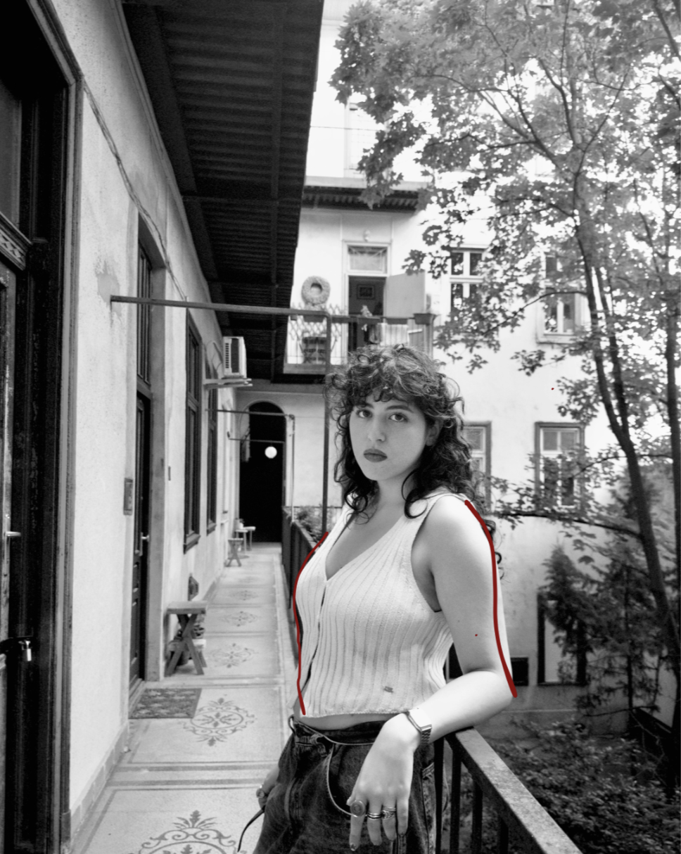 A young woman with curly hair and dark lipstick standing on a balcony in a black-and-white photo. She is wearing a sleeveless, striped top and dark pants, with rings and a watch on her left wrist. The balcony is adjacent to an old building with multiple second-story windows, some open, and a tree with leafy branches nearby.